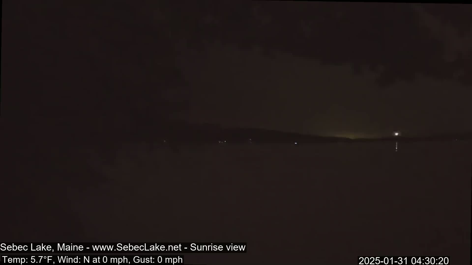 A dark, nighttime view of a calm lake with a distant, dimly lit shoreline under a cloudy sky.