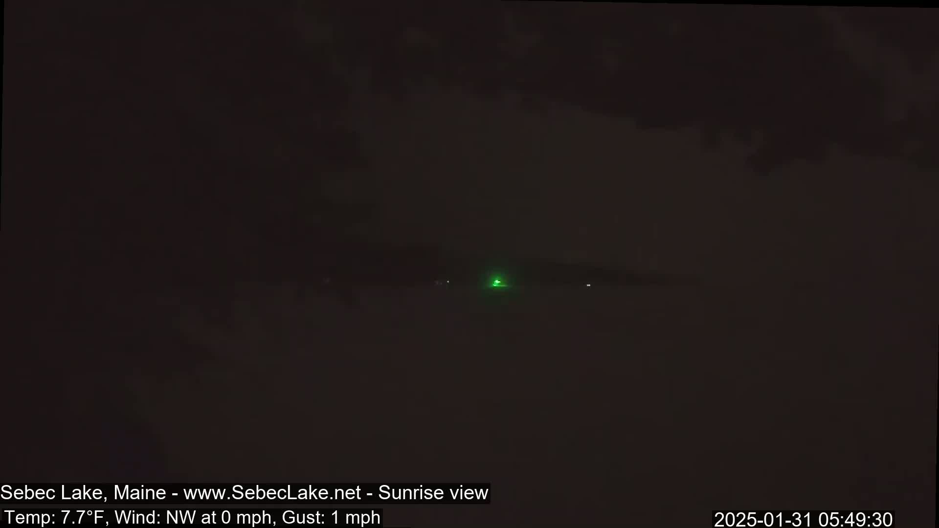 A dark, pre-dawn scene shows a body of water with a small green light visible in the distance under a mostly cloudy sky.