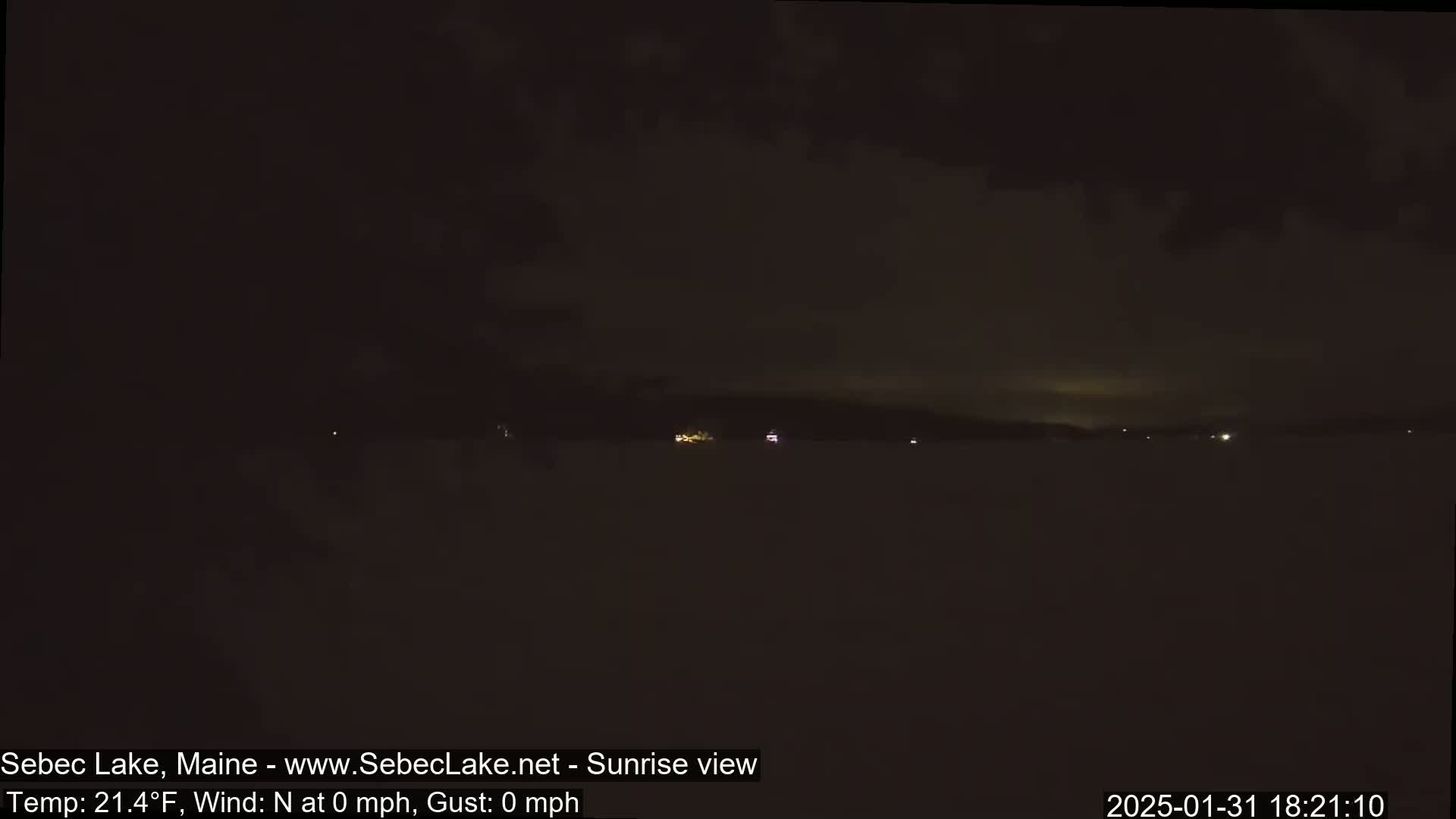A dark, nighttime view of a calm lake with a few distant lights on the far shore under a cloudy sky.