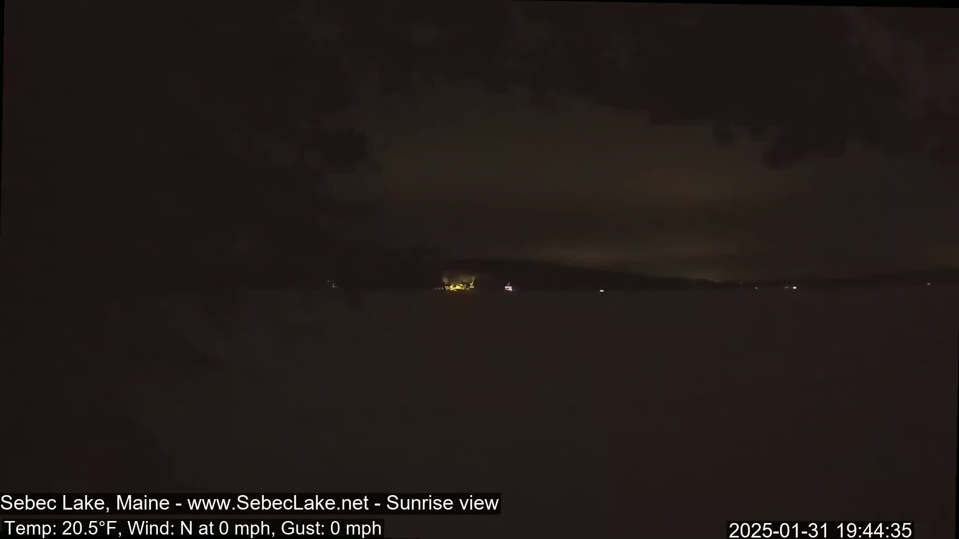 A dark, nighttime view of a calm lake with a few distant lights on the shoreline under a mostly cloudy sky.