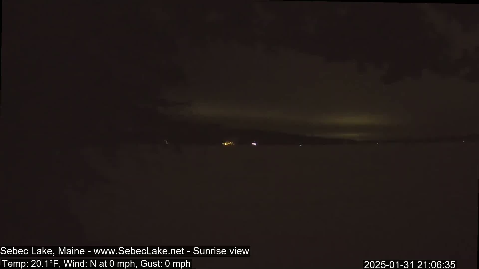 A dark, nighttime view of a snow-covered lake with a few faint lights visible in the distance under a mostly cloudy sky.