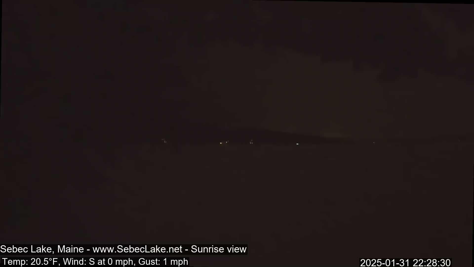 A dark, nighttime view of a lake with a few distant lights and a mostly cloudy sky, with a temperature of 20.5°F and a light southerly wind.