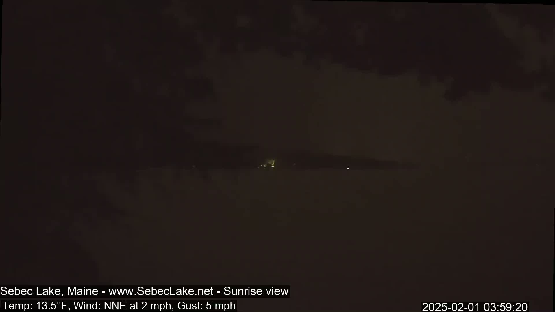 A dark, nighttime view of a lake shows a faint, distant light source on the horizon under a mostly cloudy sky.