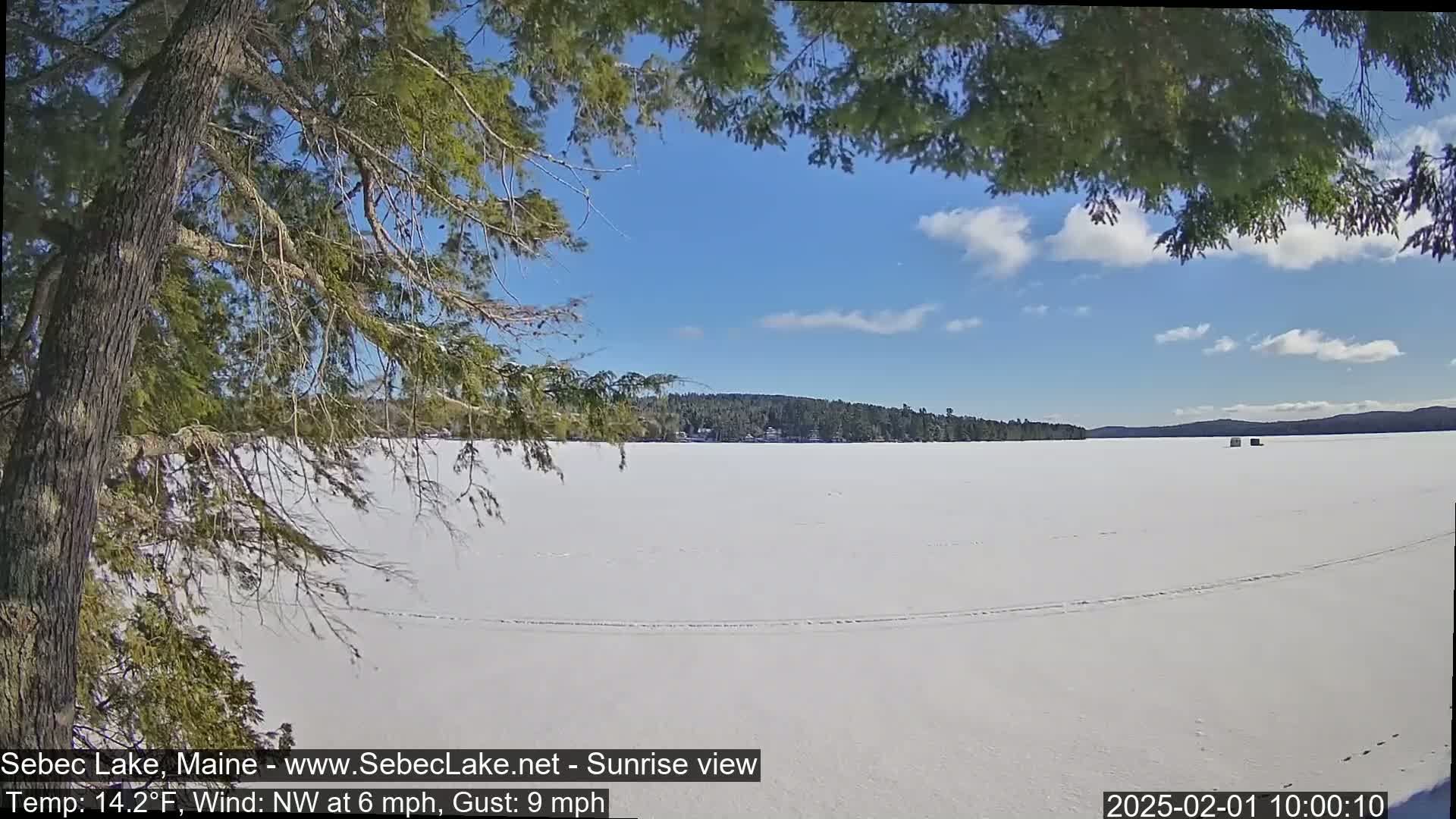 A mostly sunny day shows a frozen lake under a partly cloudy sky, viewed from behind tree branches.