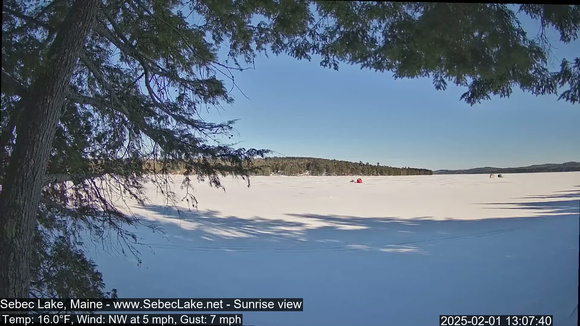 A snow-covered frozen lake is seen from a wooded shoreline under a clear blue sky, with a few ice fishing shelters visible in the distance.