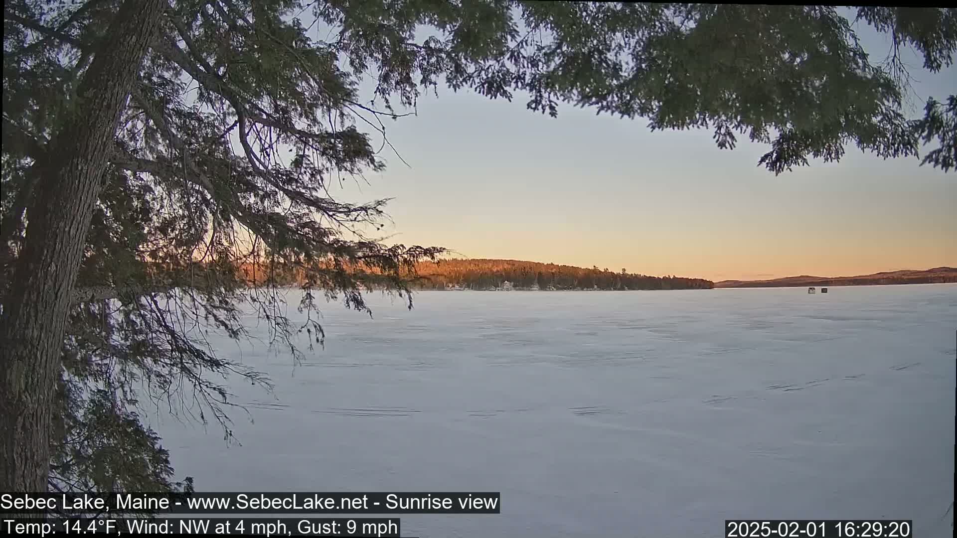 A frozen lake at sunset is viewed through the branches of a tree, with a light northwest wind.