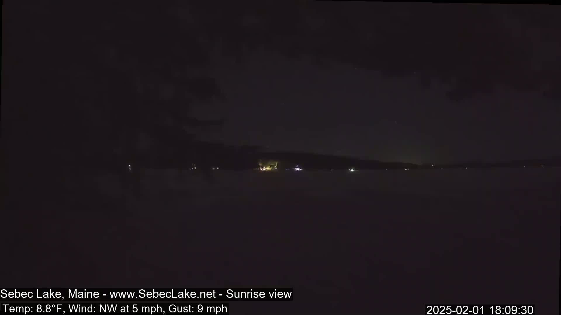 A dark, nighttime view of a lake with a few distant lights across the water, under a mostly clear sky, with a northwest wind at 5 mph and gusts up to 9 mph.