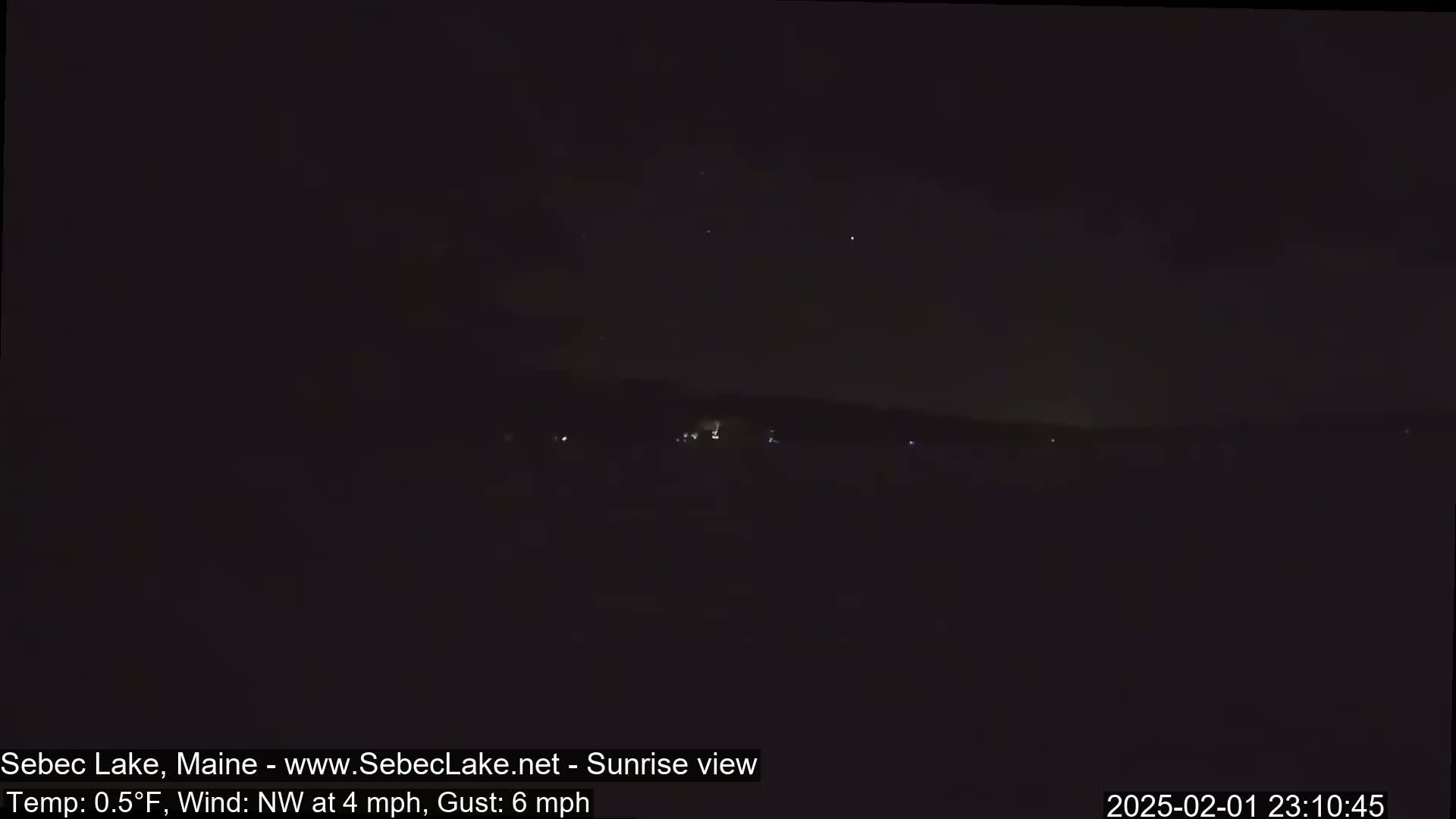 A dark, nighttime view of a landmass with a few faint lights visible in the distance, under a mostly clear sky with a northwest wind.