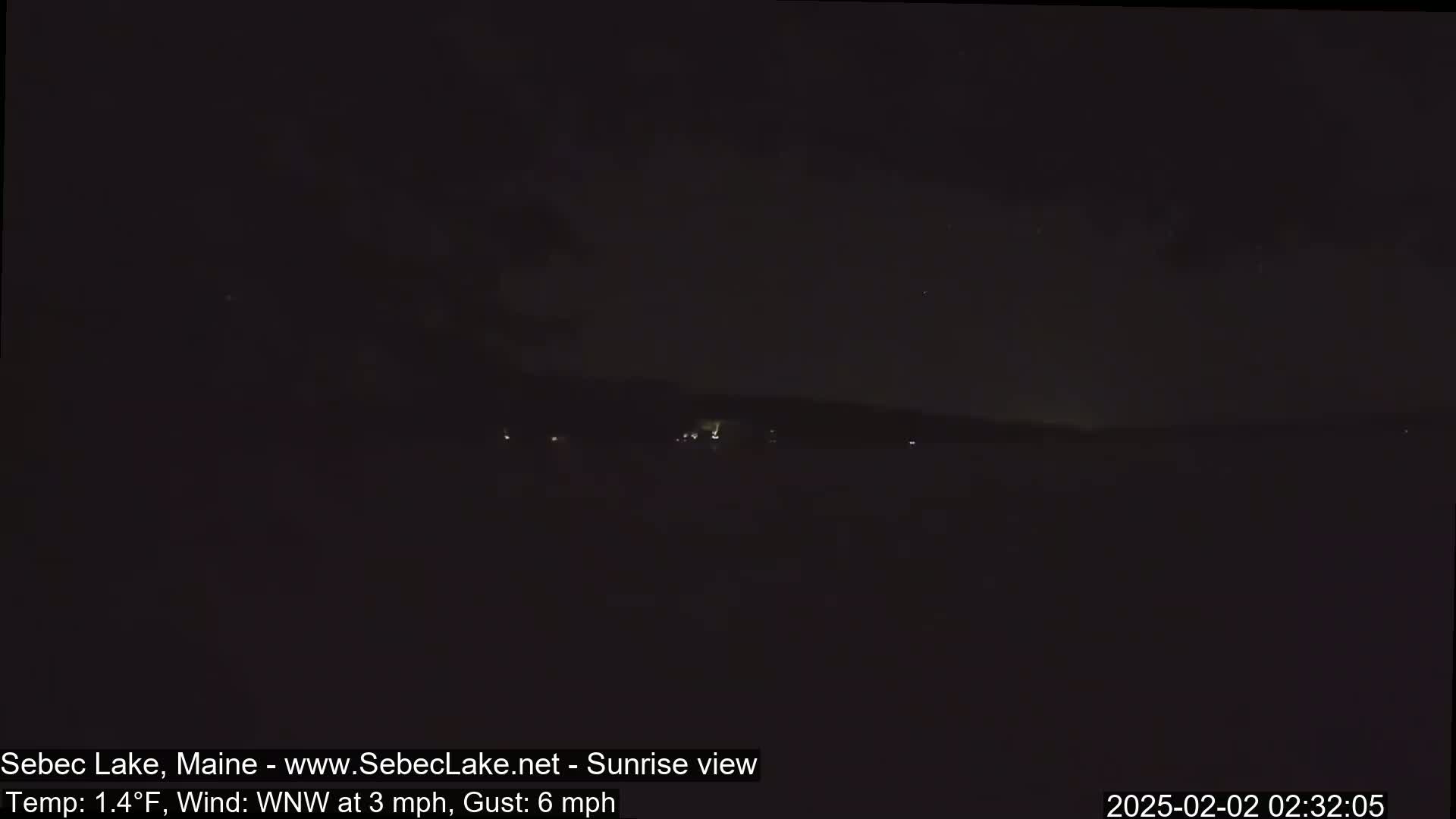 A dark, nighttime view of a lake with a distant shoreline and a few faint lights visible under a cloudy sky, with a temperature of 1.4°F and a wind from the west-northwest at 3 mph, gusting to 6 mph.