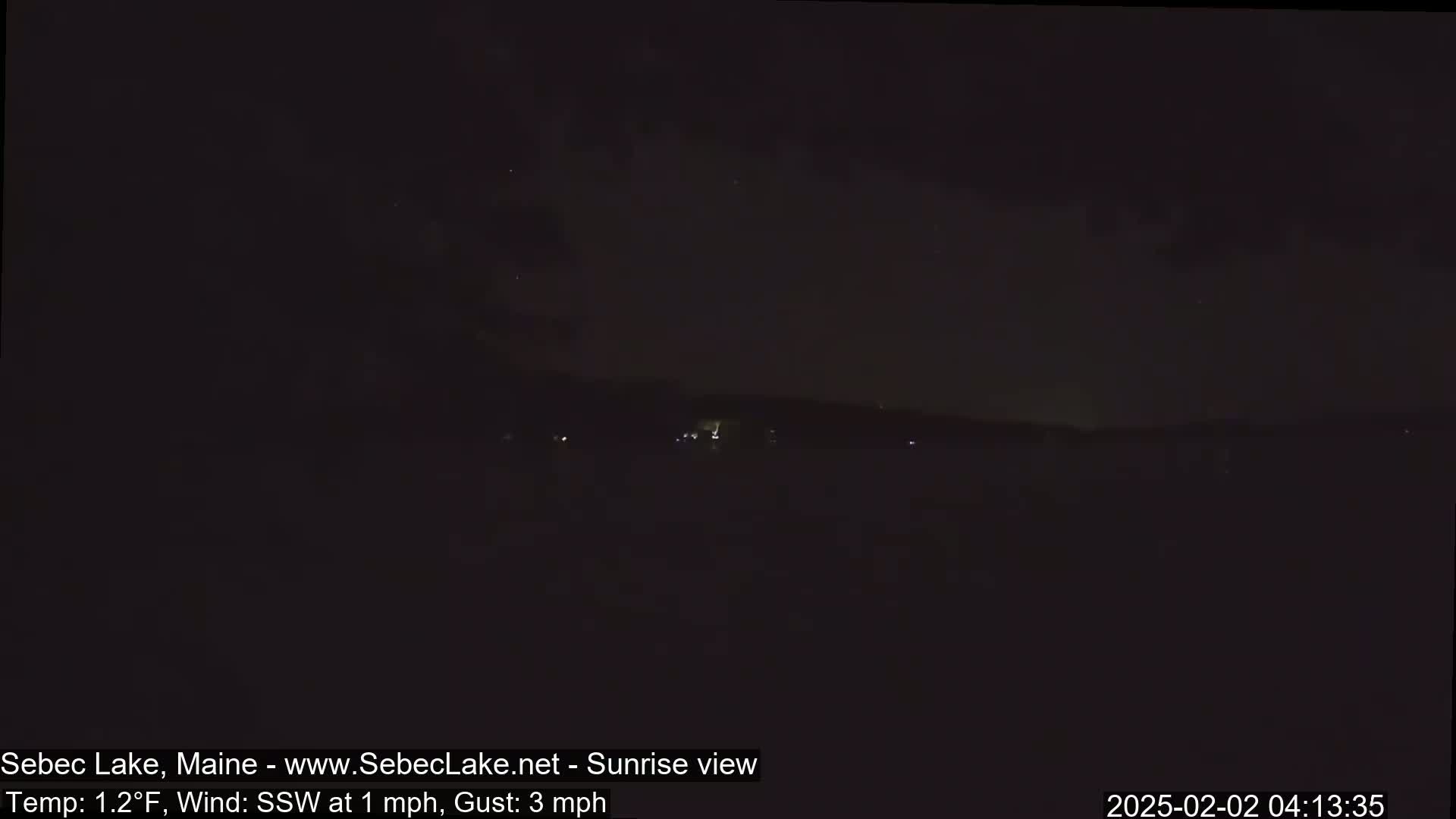 A dark, nighttime view of a lake with a distant shoreline under a slightly cloudy sky.