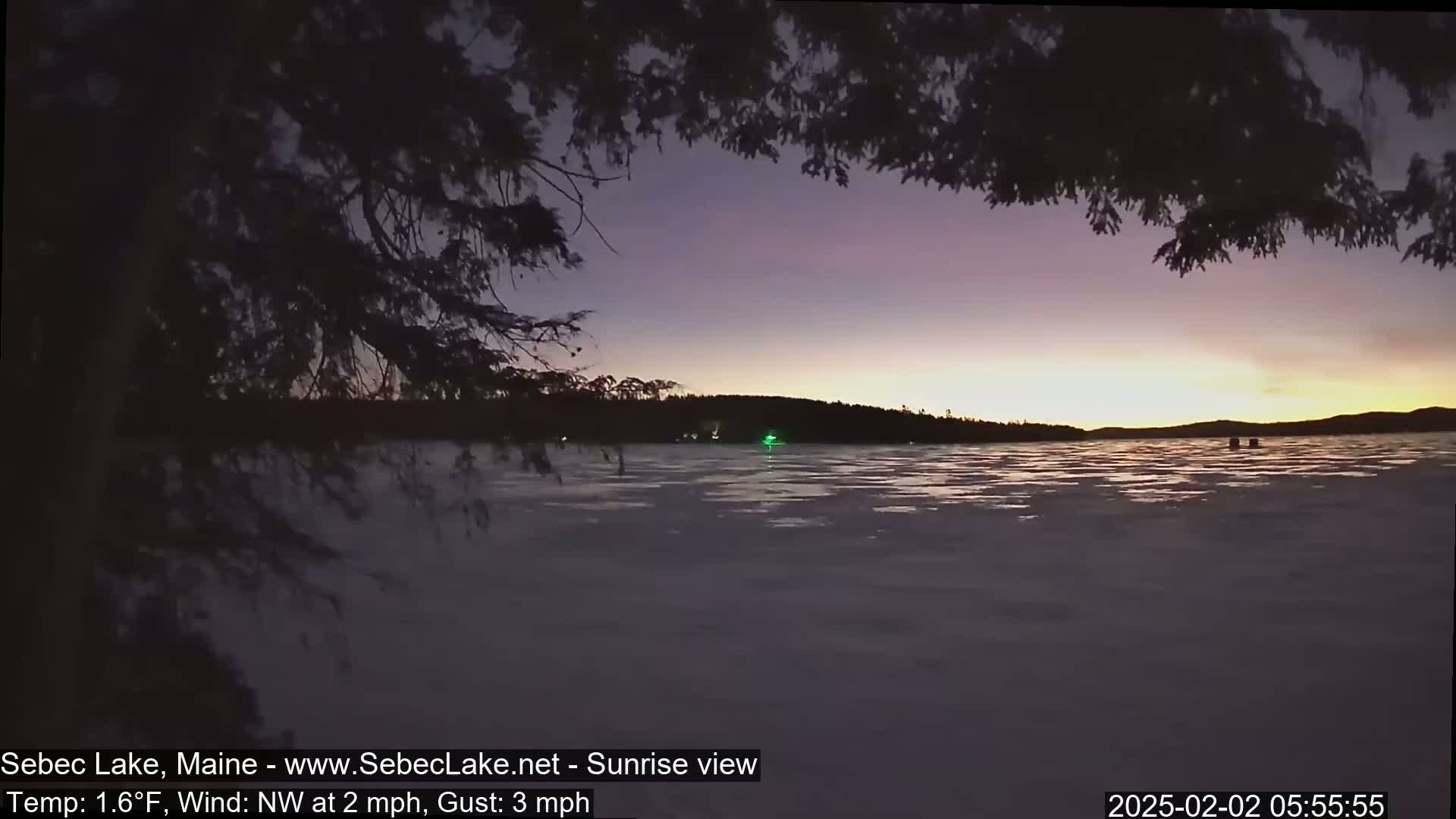 A partially frozen lake at sunrise is viewed from behind a tree's branches, with a calm, light wind blowing from the northwest.