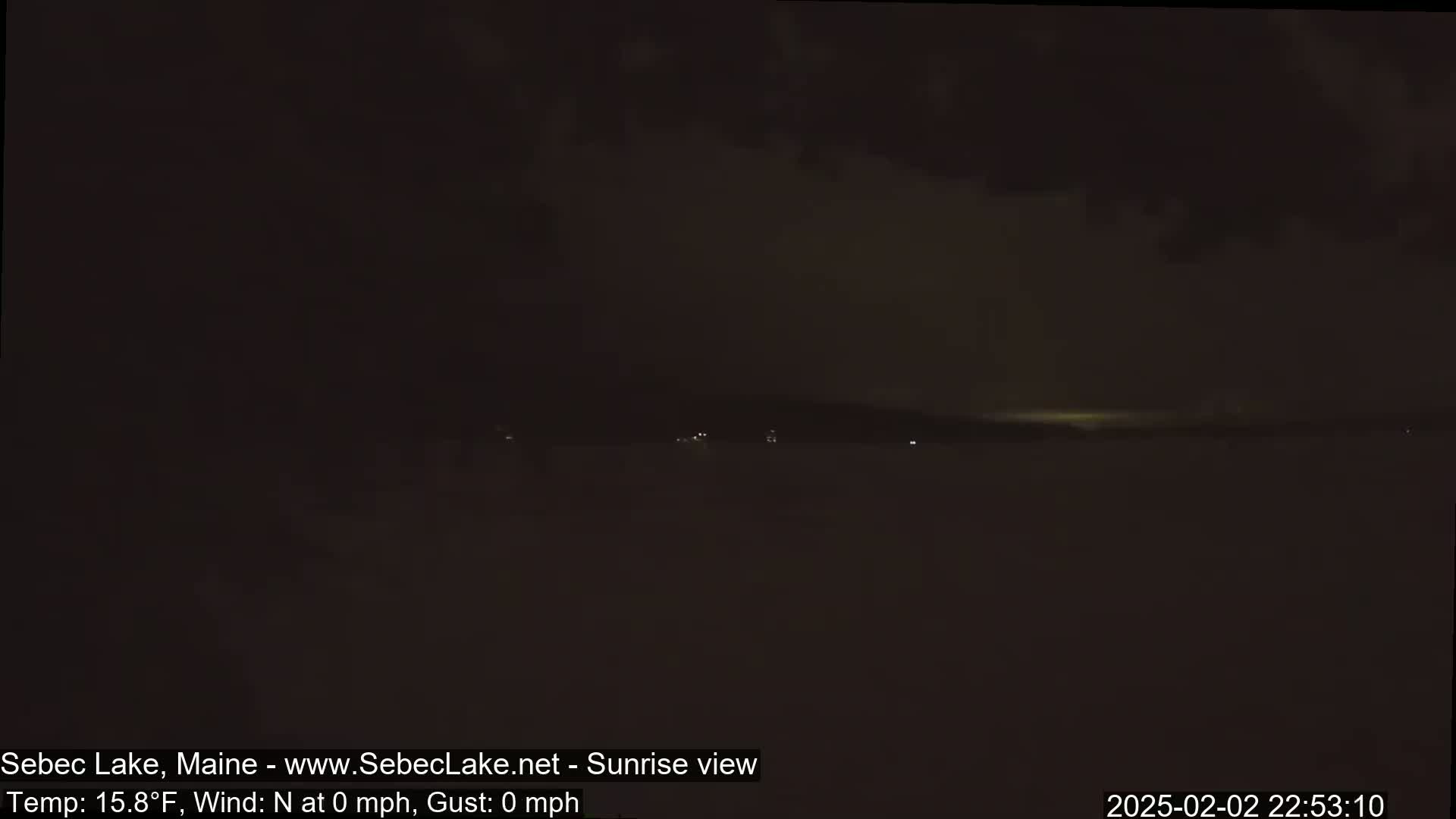 A dark, nighttime view of a lake with a faint, distant glow on the horizon, under calm, cold conditions.