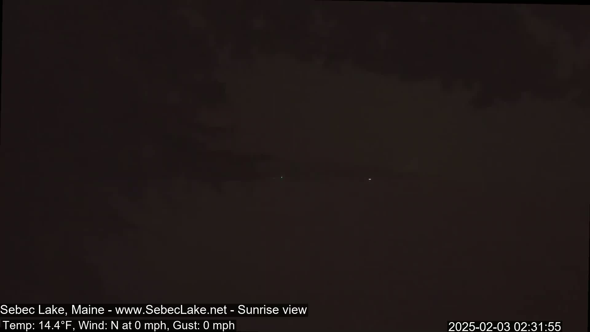 A dark, pre-dawn view of a lake shows a very faint horizon line under a mostly cloudy sky, with the temperature at 14.4°F and no wind.