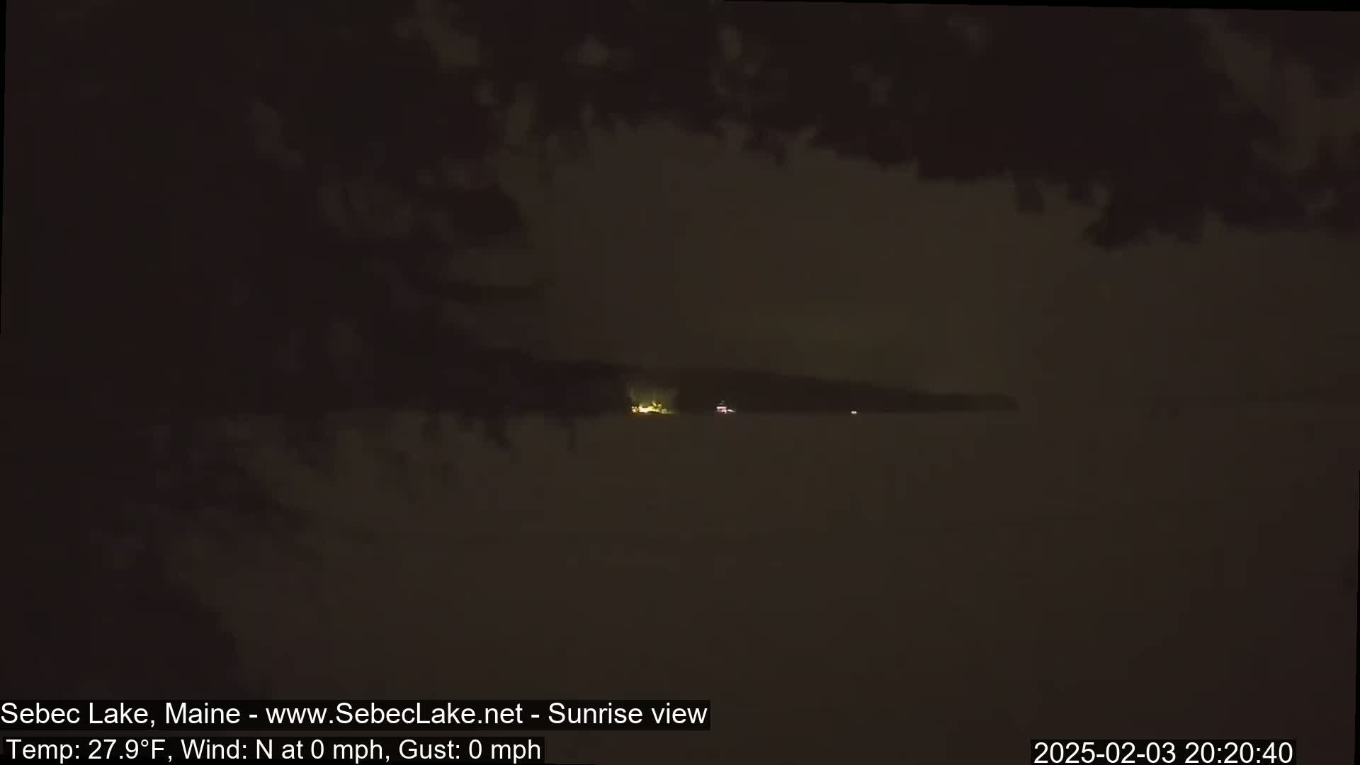 A dark, nighttime view of a lake shows a distant shoreline with faint lights under a cloudy sky.