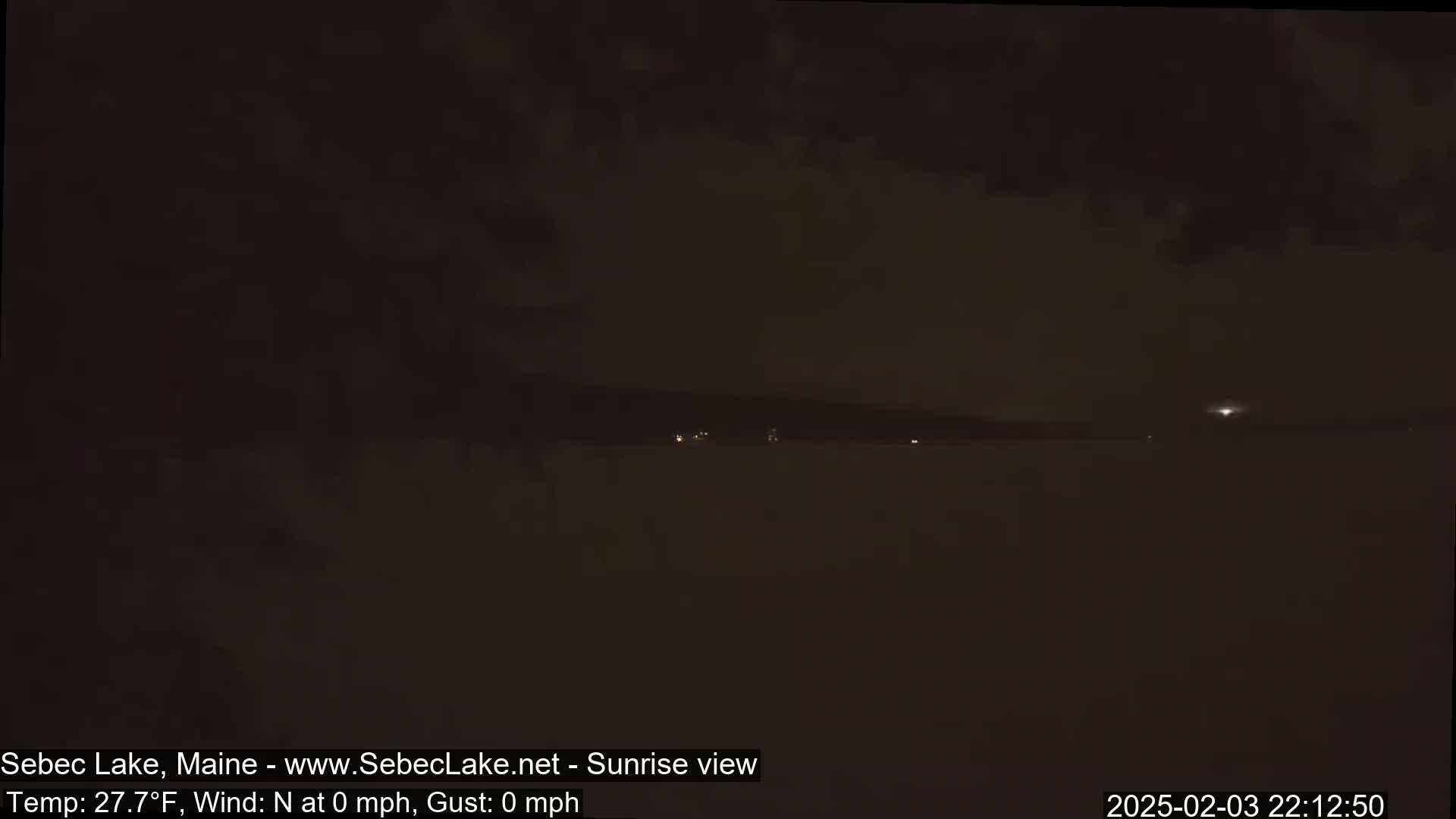 A dark, nighttime view of a lake with a distant shoreline showing a few faint lights under a mostly cloudy sky.