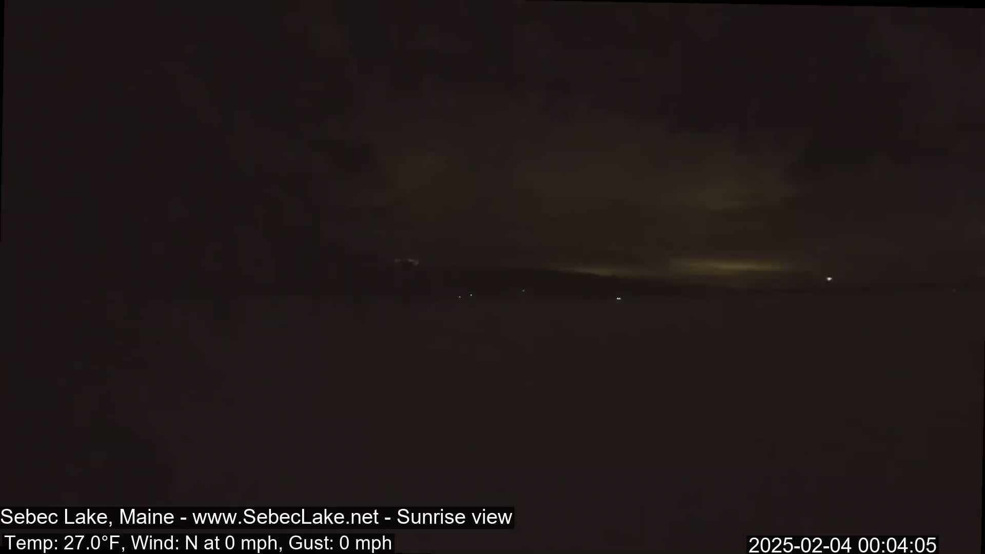 A dark, nighttime scene shows a still body of water under a mostly cloudy sky, with a few faint lights visible in the distance.