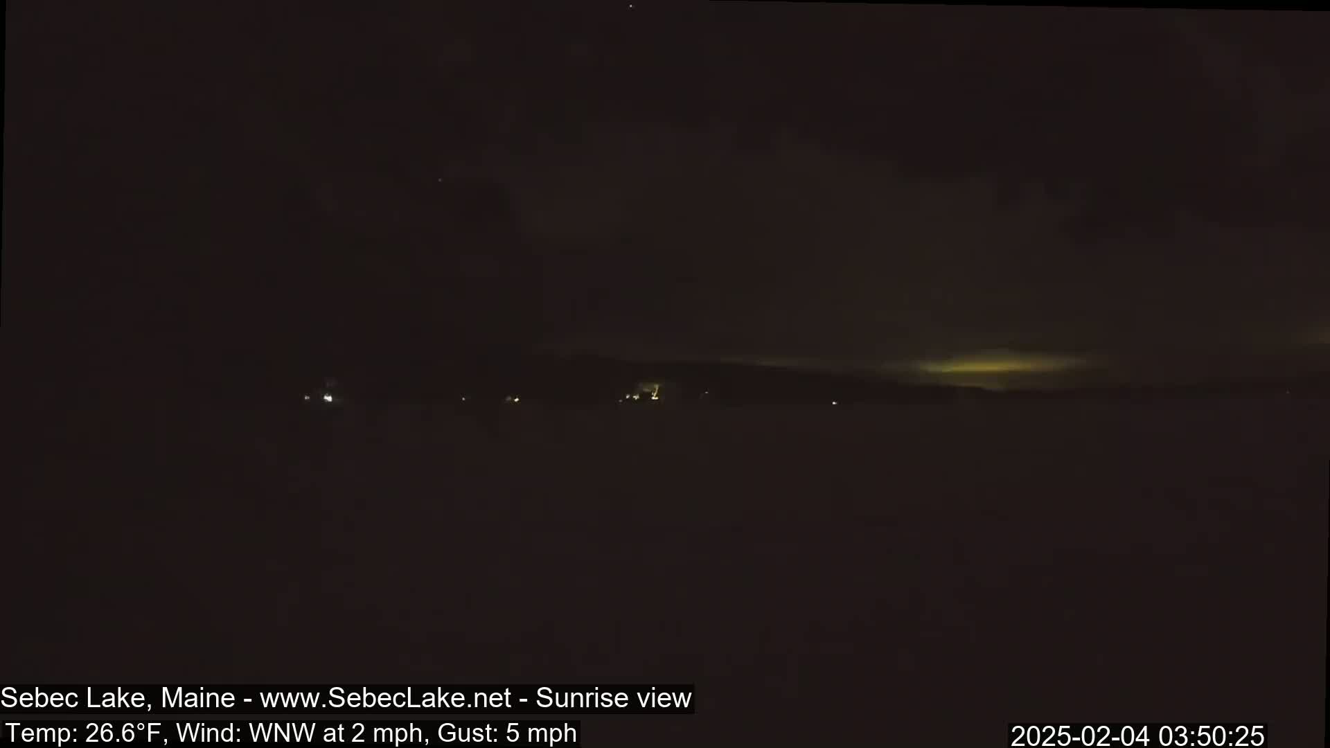 A dark, nighttime landscape shows a faint, yellowish glow on the horizon under a mostly cloudy sky, with a light wind.
