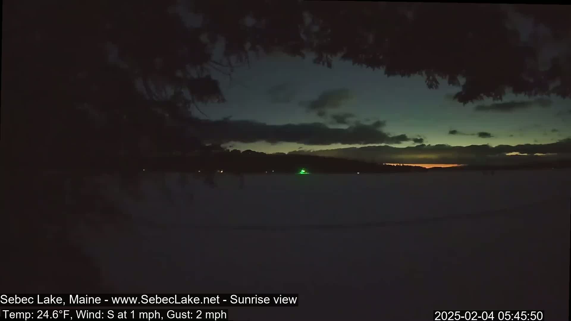 A dark, pre-dawn view of a snow-covered lake with a barely visible sunrise, under a mostly cloudy sky with a light southerly wind.