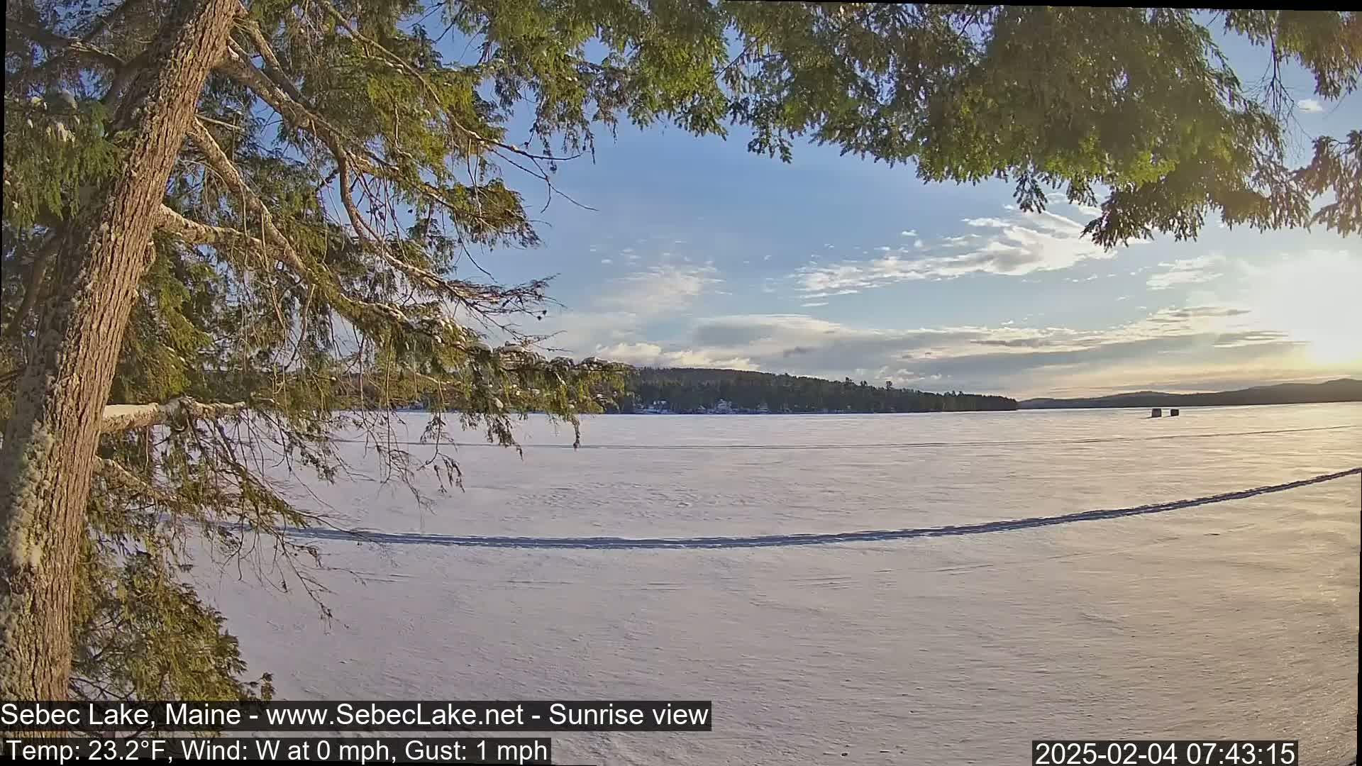 A snow-covered frozen lake is viewed from under the branches of a snow-dusted evergreen tree, with a partly cloudy sky and a calm sunrise.