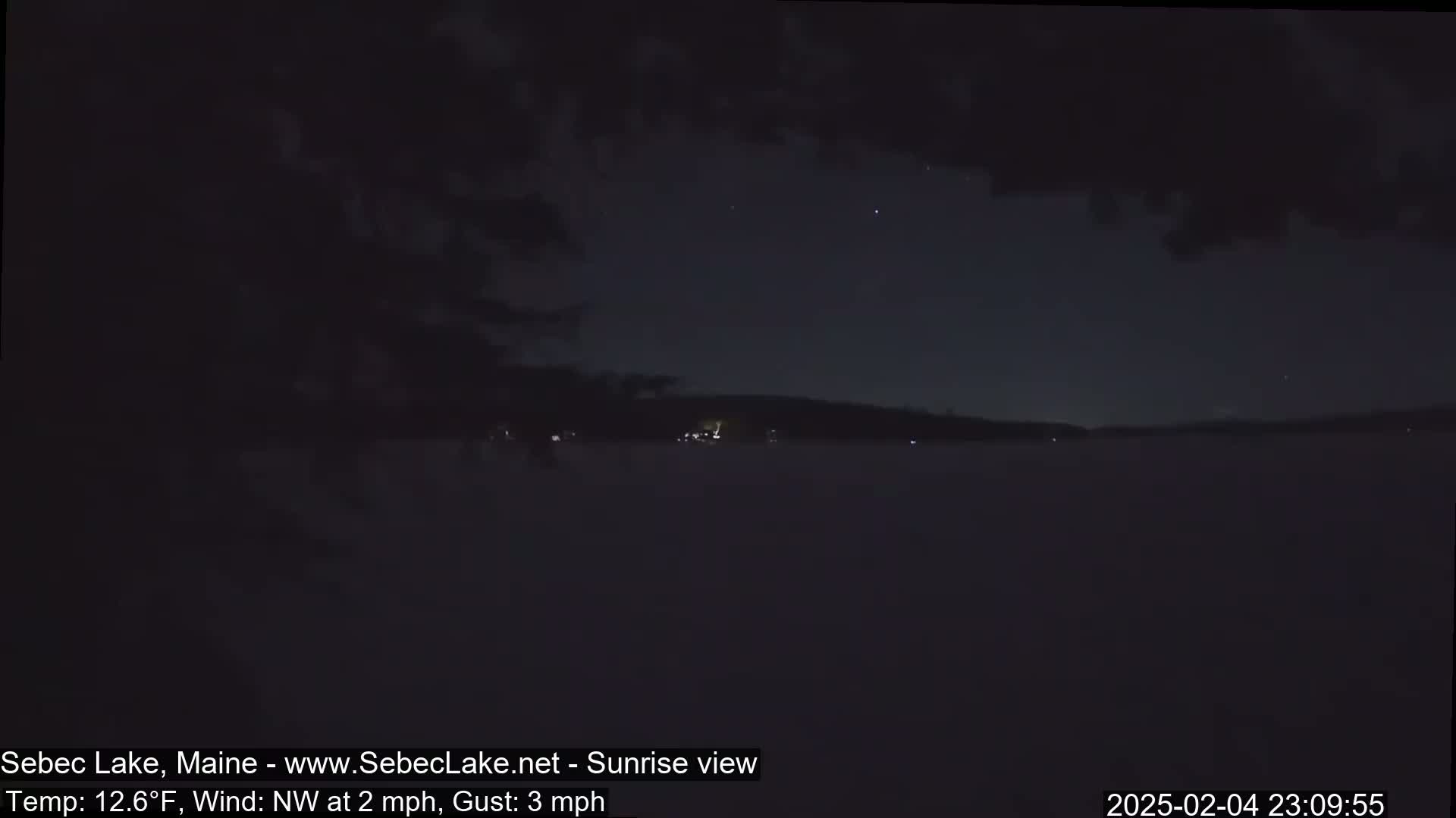 A dark, snowy landscape at night shows a lake under a mostly cloudy sky with a few stars visible.