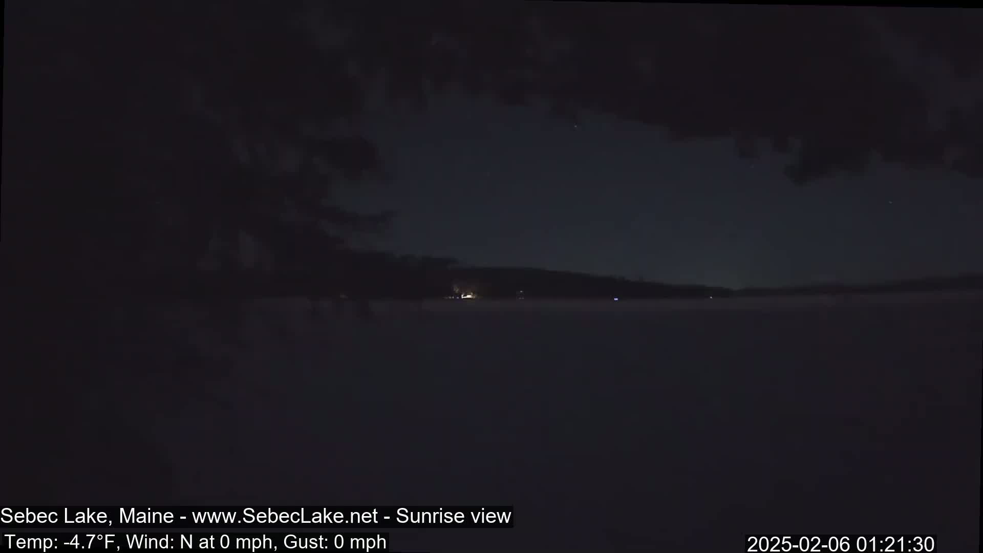 A dark, nighttime view of a calm lake with a distant shoreline under a cloudy sky.