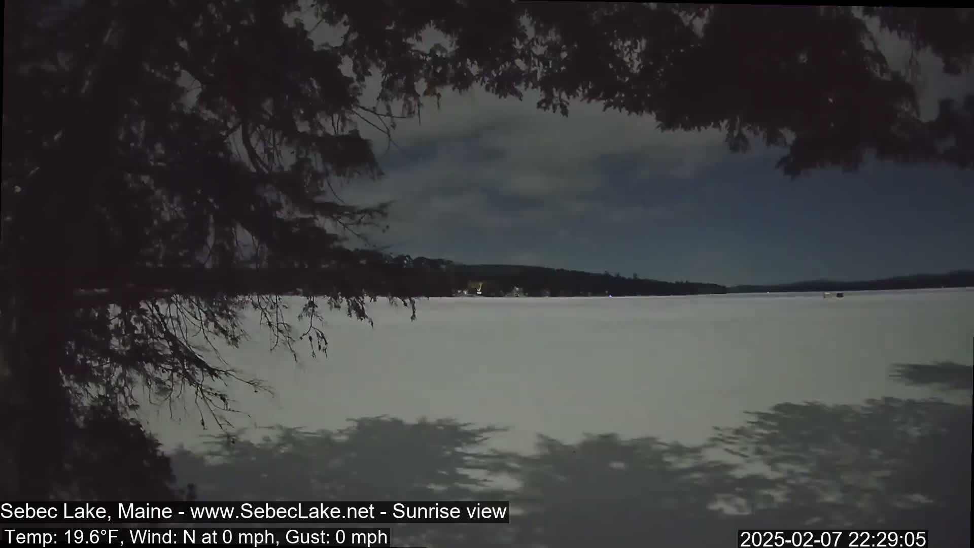 A mostly cloudy, calm night scene shows a frozen lake under a dark sky, viewed from behind tree branches casting shadows on the snow-covered ice.