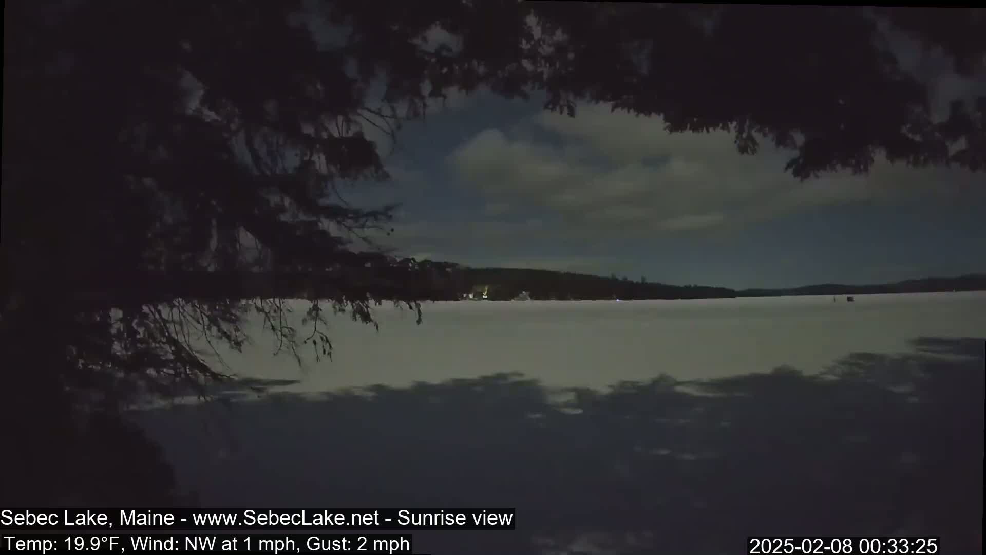 A frozen lake is visible under a mostly cloudy night sky, with tree branches partially obscuring the view.