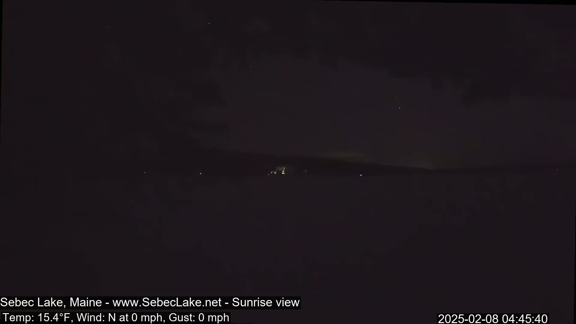 A dark, nighttime view of a lake shows a distant, faintly lit shoreline under a mostly clear, starlit sky.