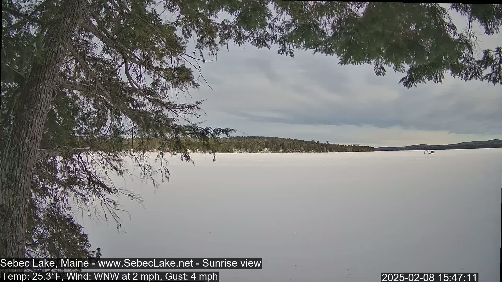 A partially obscured view of a snow-covered frozen lake under a cloudy sky shows a distant treeline and a few small structures on the ice.