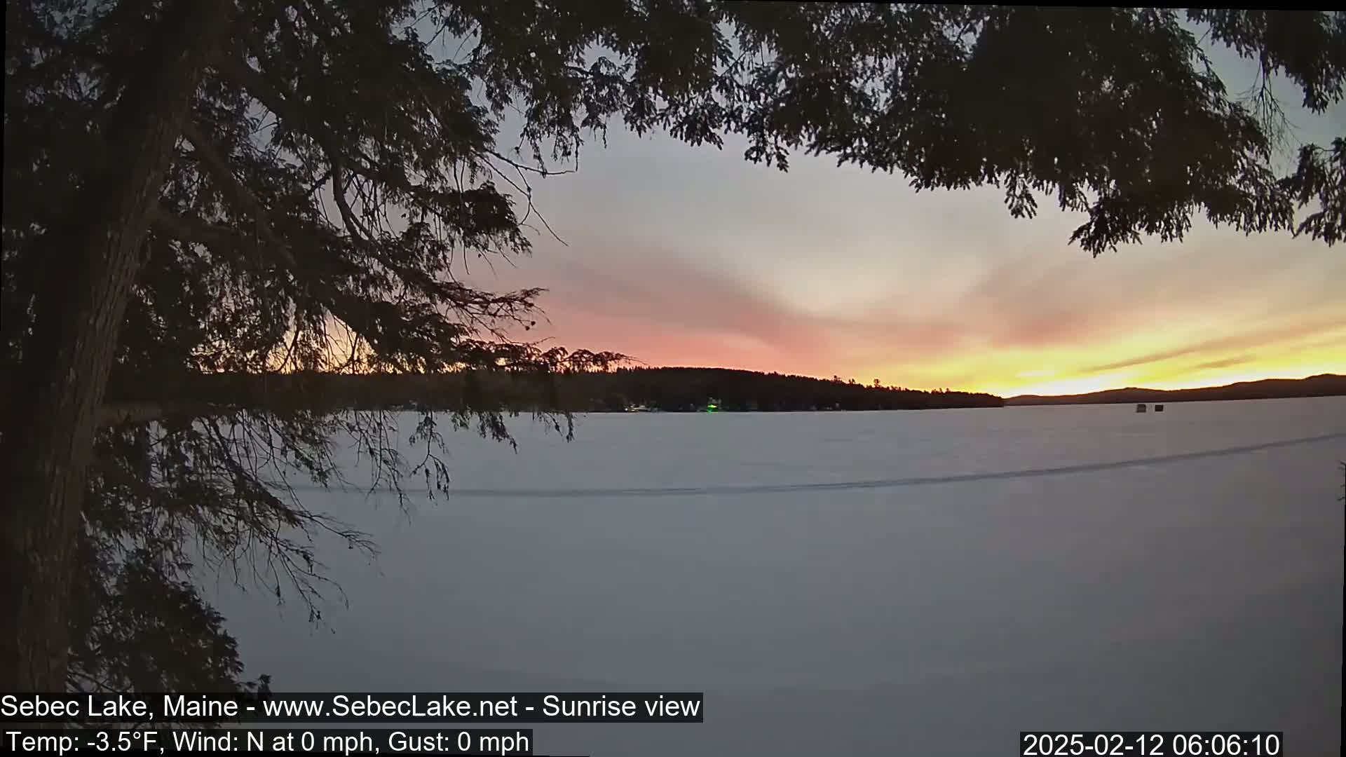 A sunrise with pink and orange hues is visible over a frozen lake, partially obscured by dark tree branches in the foreground, under calm, cold conditions.