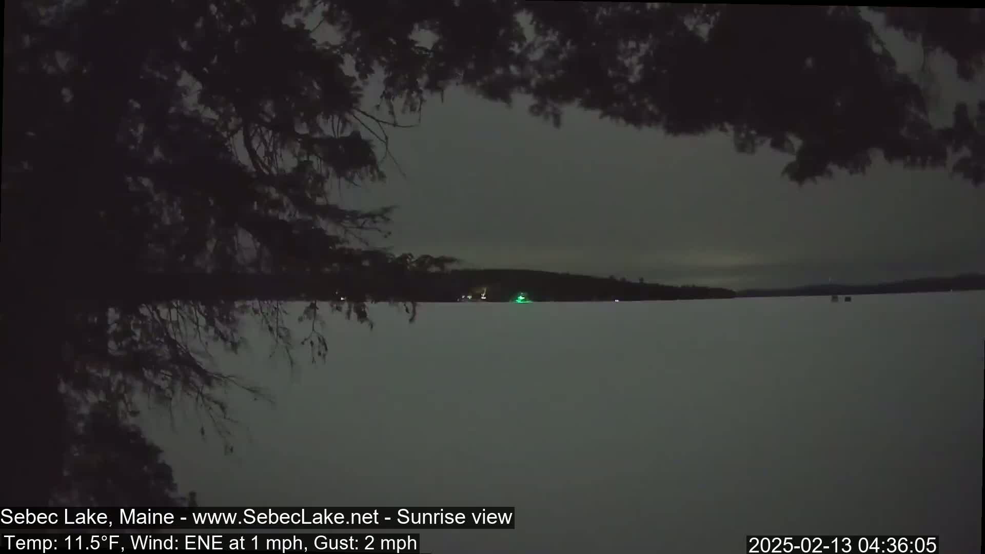 A dark, pre-dawn view of a snow-covered lake with a dark treeline in the background, under a gray sky with a light east-northeast wind.