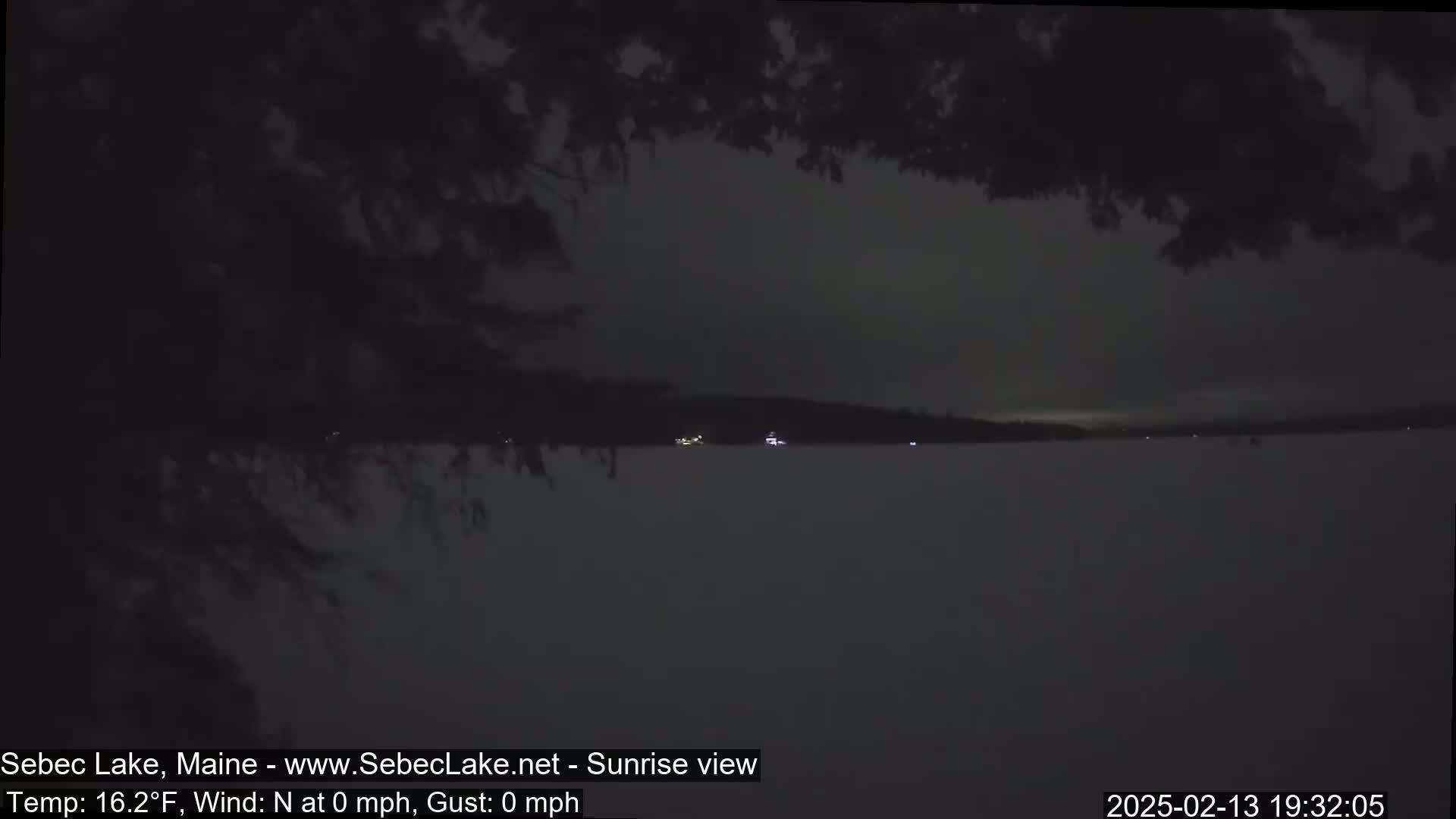 A dark, nighttime view of a snow-covered lake with a distant, dimly lit shoreline under a mostly cloudy sky.