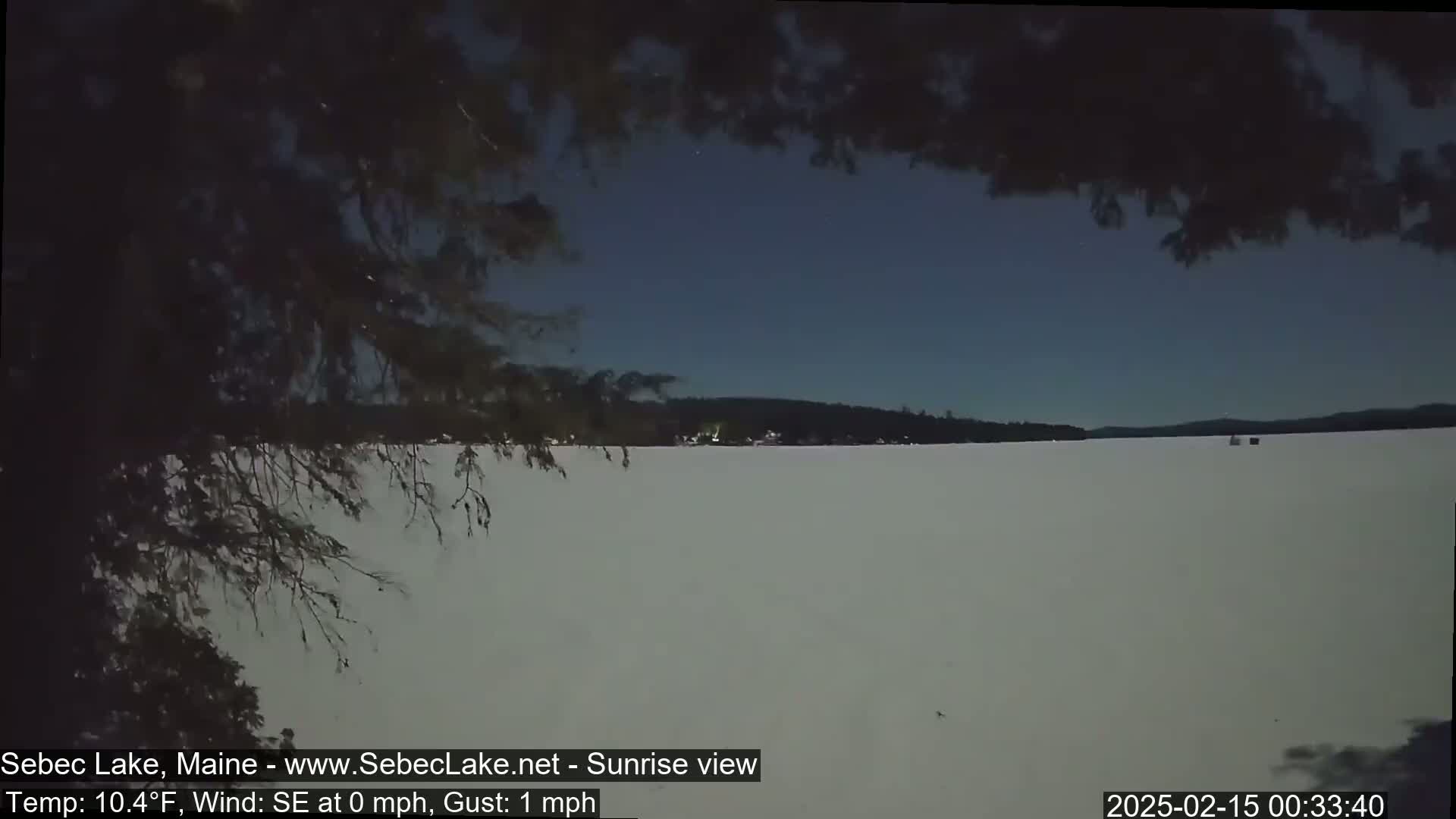 A frozen lake is visible at sunrise under a clear, dark blue sky, partially obscured by overhanging tree branches.
