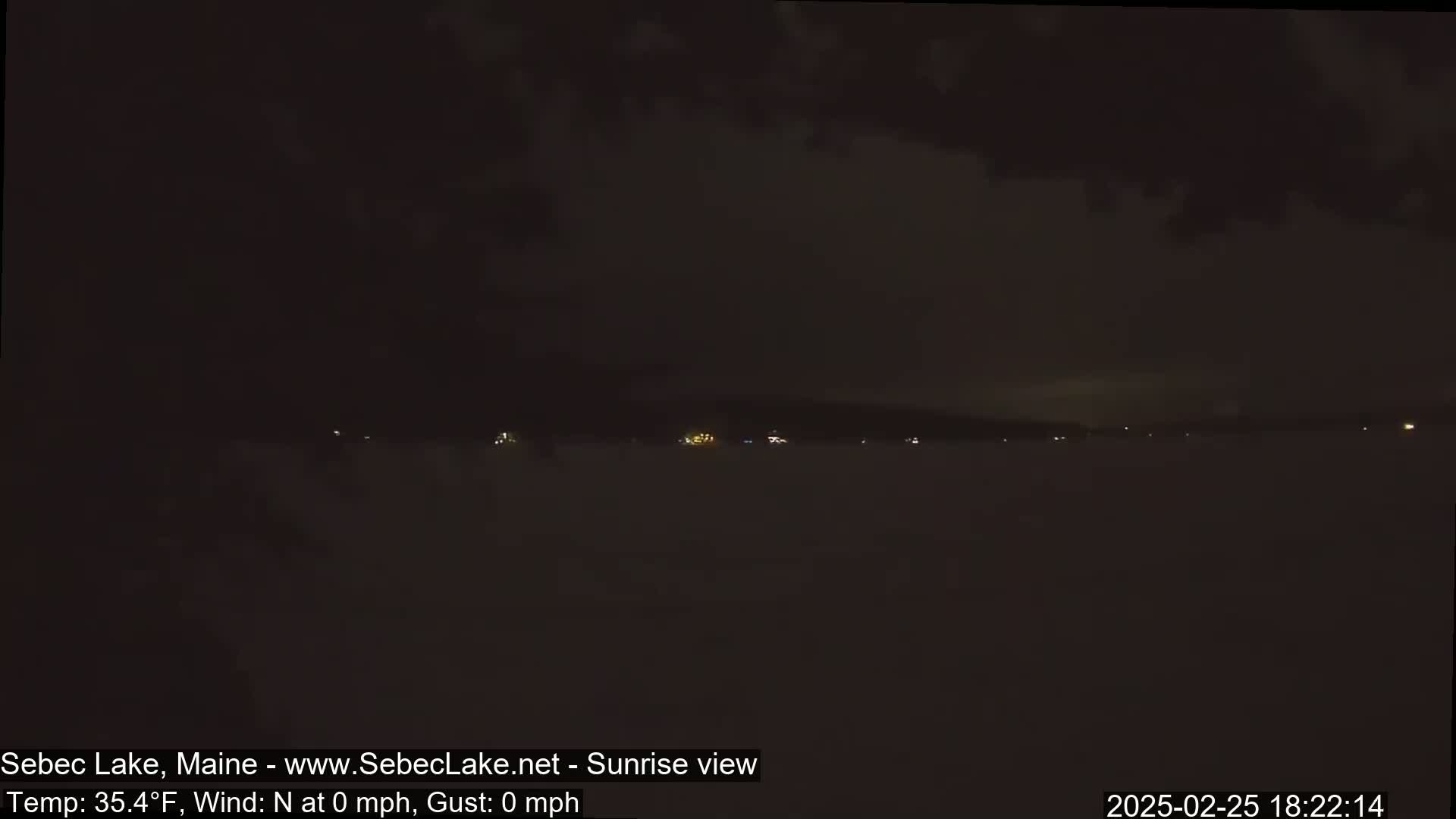 A dark, nighttime view of a lake shows a distant shoreline with faint lights under a cloudy sky.