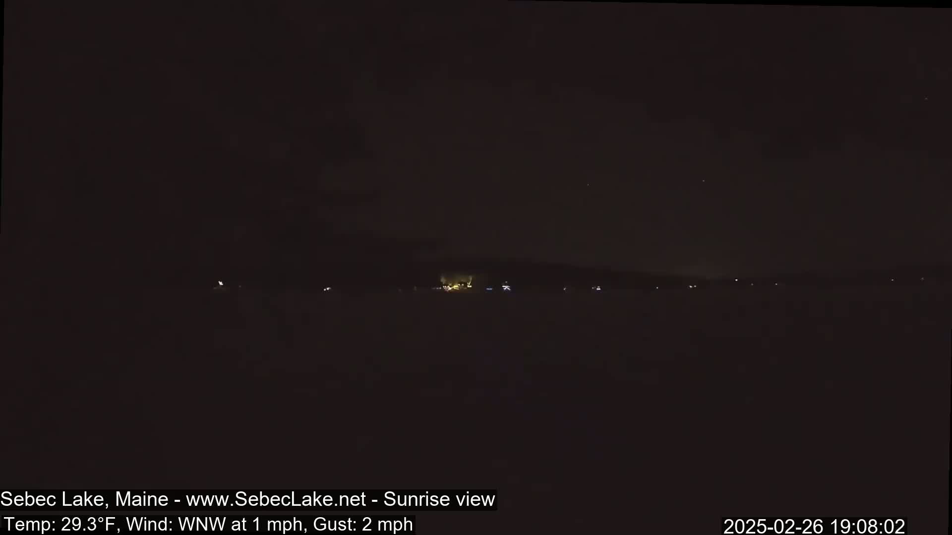 A nighttime, low-light view of a dark landscape with a few distant lights, under a dark sky with a slight wind.