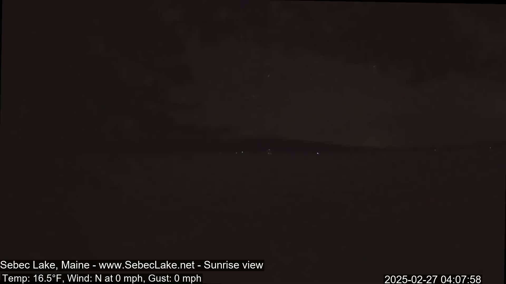 A dark, nighttime landscape shows a distant shoreline with a few faint lights under a mostly clear sky.