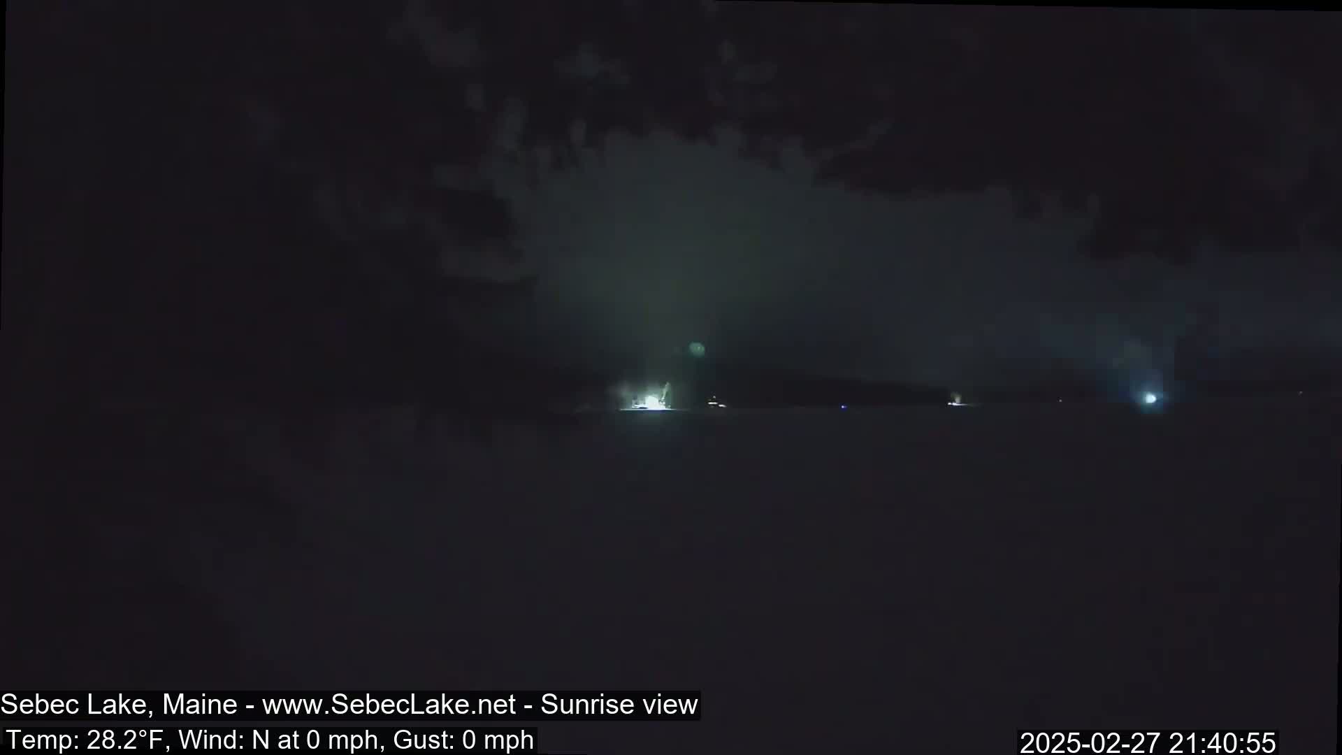 A nighttime, calm view of a dark lake with faint lights visible in the distance.