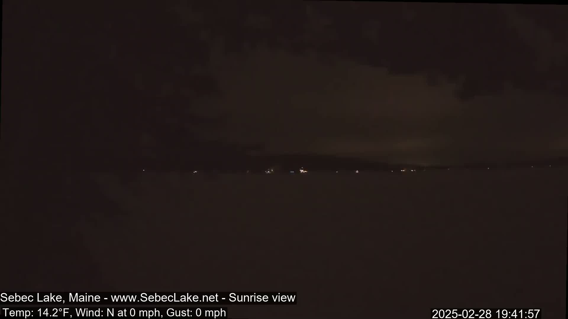 A dark, nighttime view of a lake with a few distant lights visible along the far shore under a mostly cloudy sky.
