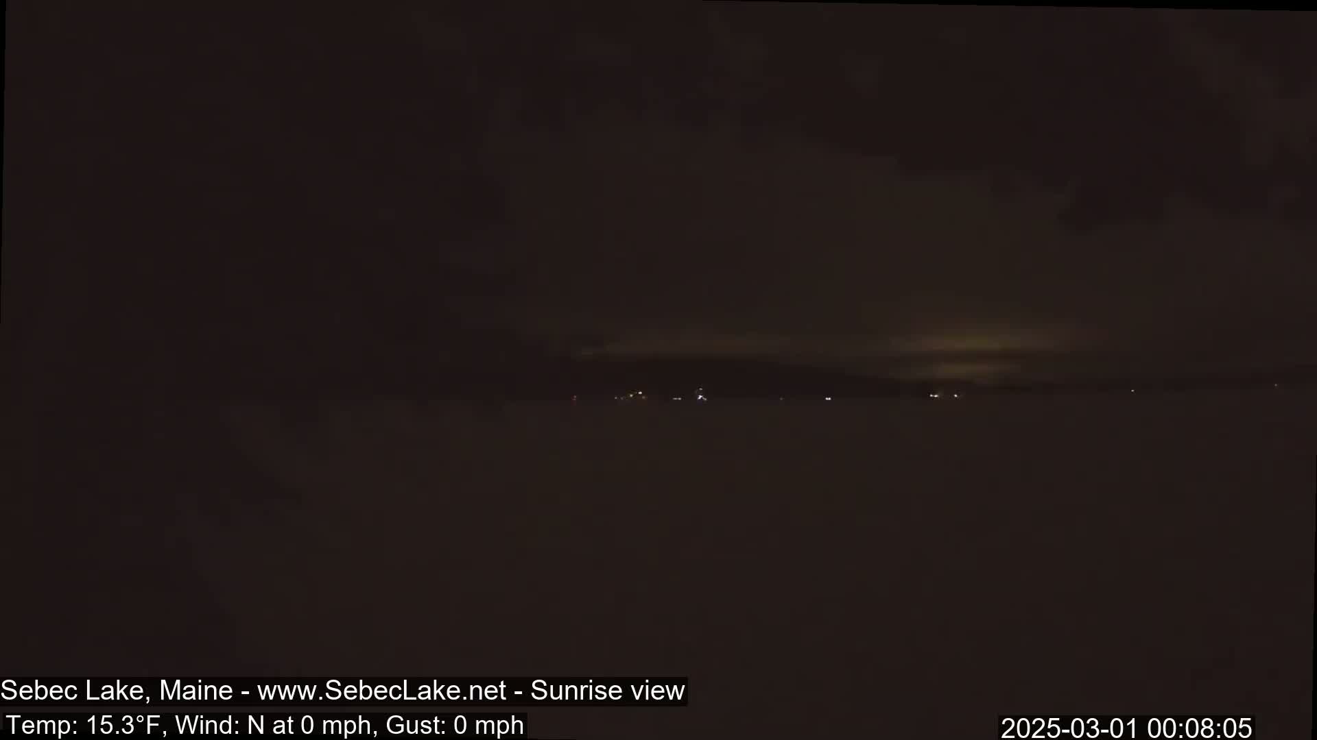 A dark, nighttime view of a snow-covered landscape with a few distant lights visible under a mostly cloudy sky.