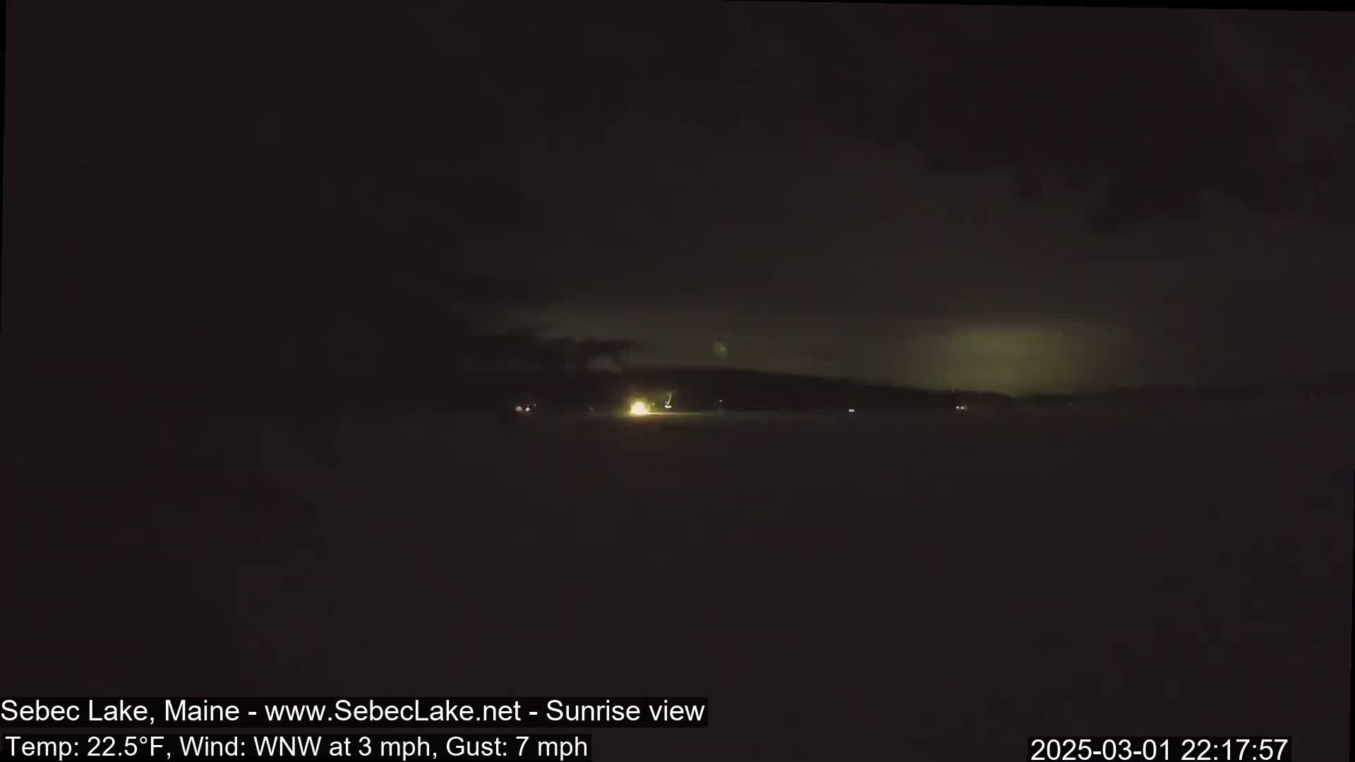 A nighttime long shot shows a dark, possibly snowy, landscape with faint lights in the distance under a cloudy sky.