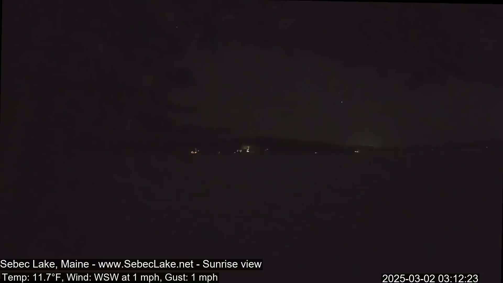 A dark, nighttime view of a lake with a few distant lights visible on the far shore, under a clear sky with a faint WSW wind of 1 mph.