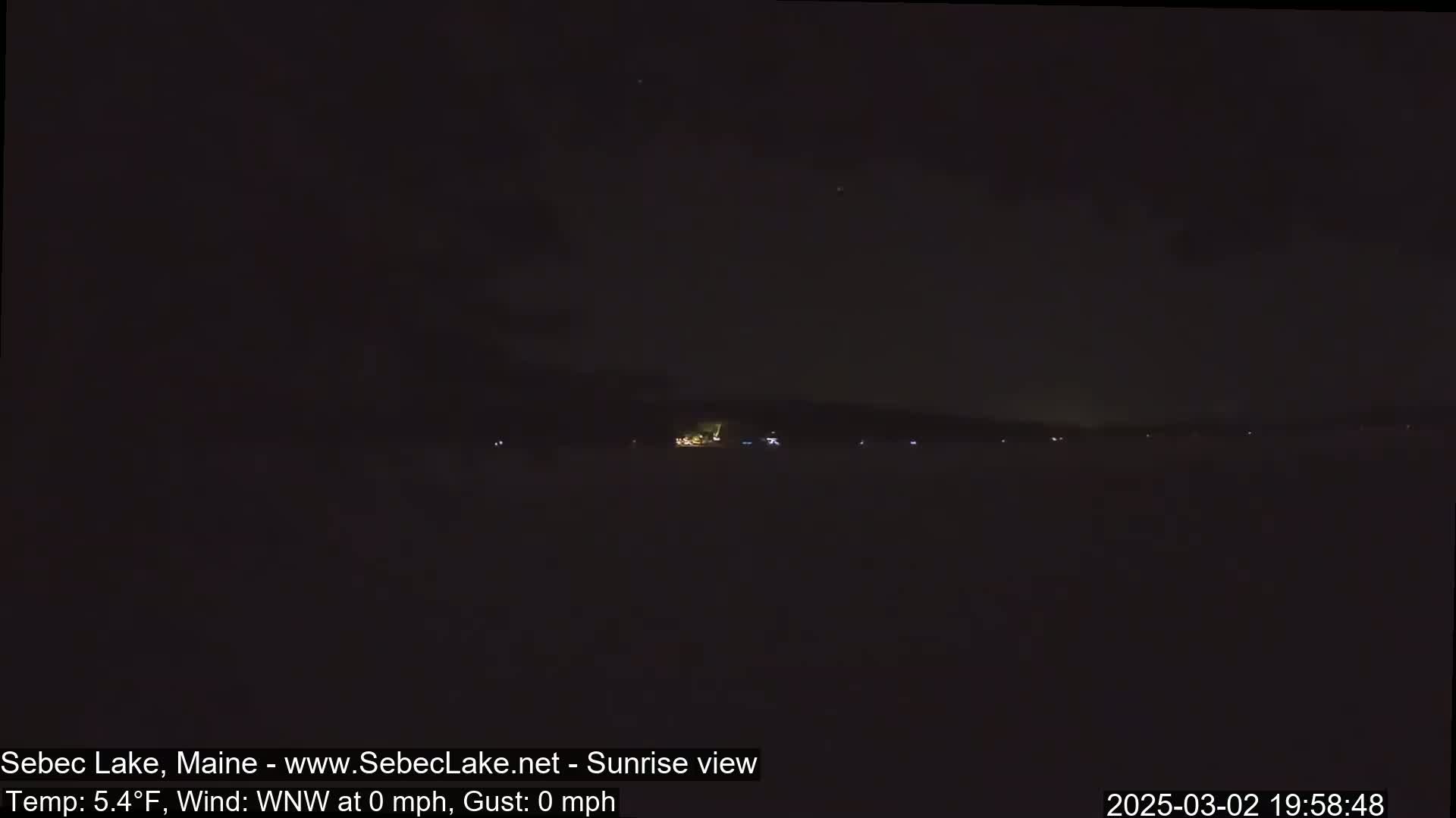 A dark, nighttime view of a lake with a distant shoreline showing a few faint lights under a clear, starlit sky.