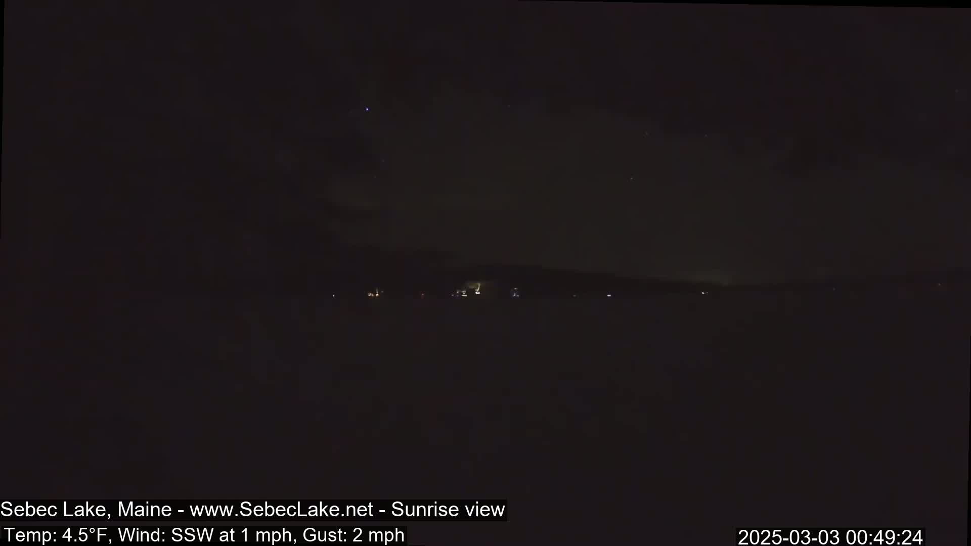 A nighttime long shot of a dark lake with a few faint lights on the far shore under a mostly clear, starry sky.
