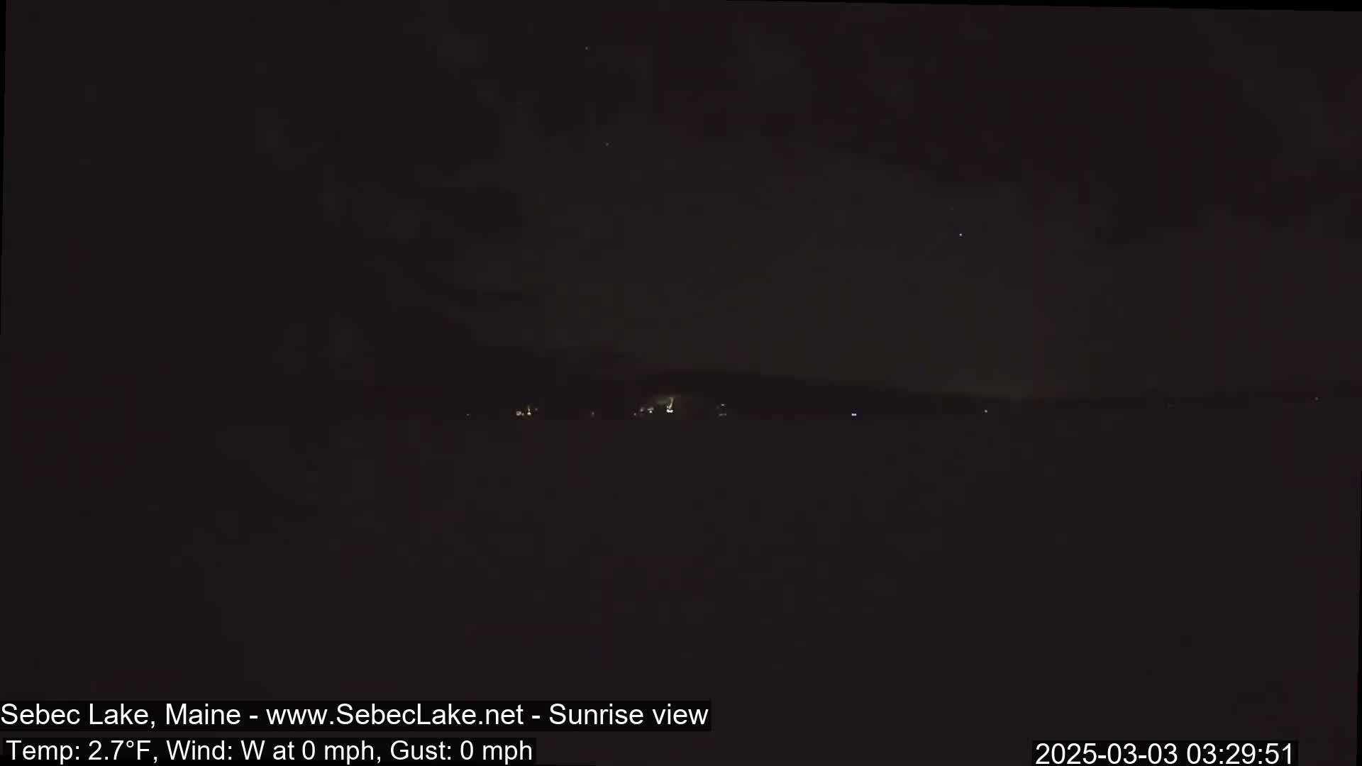 A dark, nighttime view of a calm lake with a distant shoreline, under a mostly clear sky.