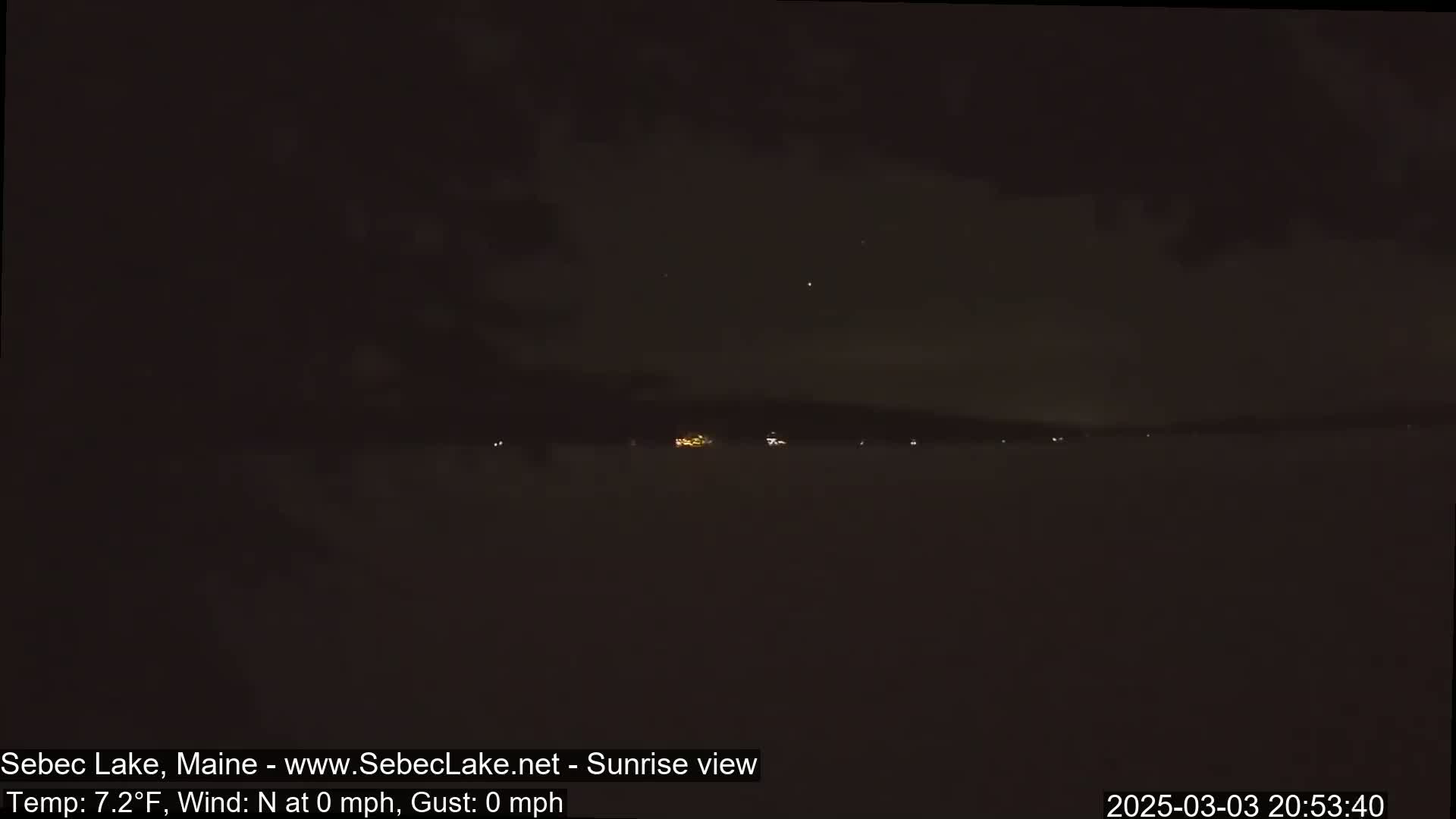 A nighttime view of a calm lake with a dark sky and a few distant lights on the far shore.