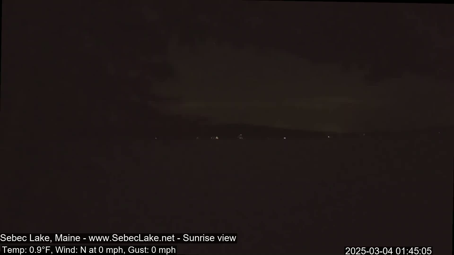 A dark, nighttime view of a lake with a few distant lights, under a mostly cloudy sky and calm, cold conditions.