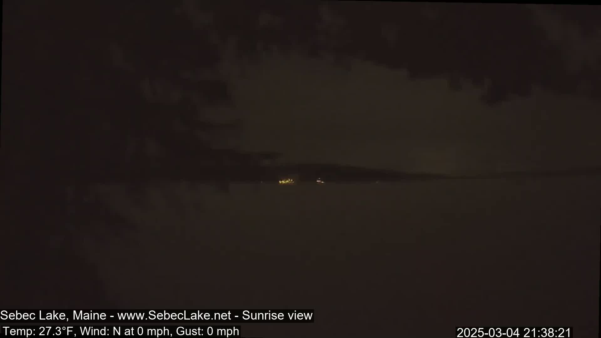A dark, nighttime view of a lake shows a distant shoreline with faint lights under a mostly cloudy sky.