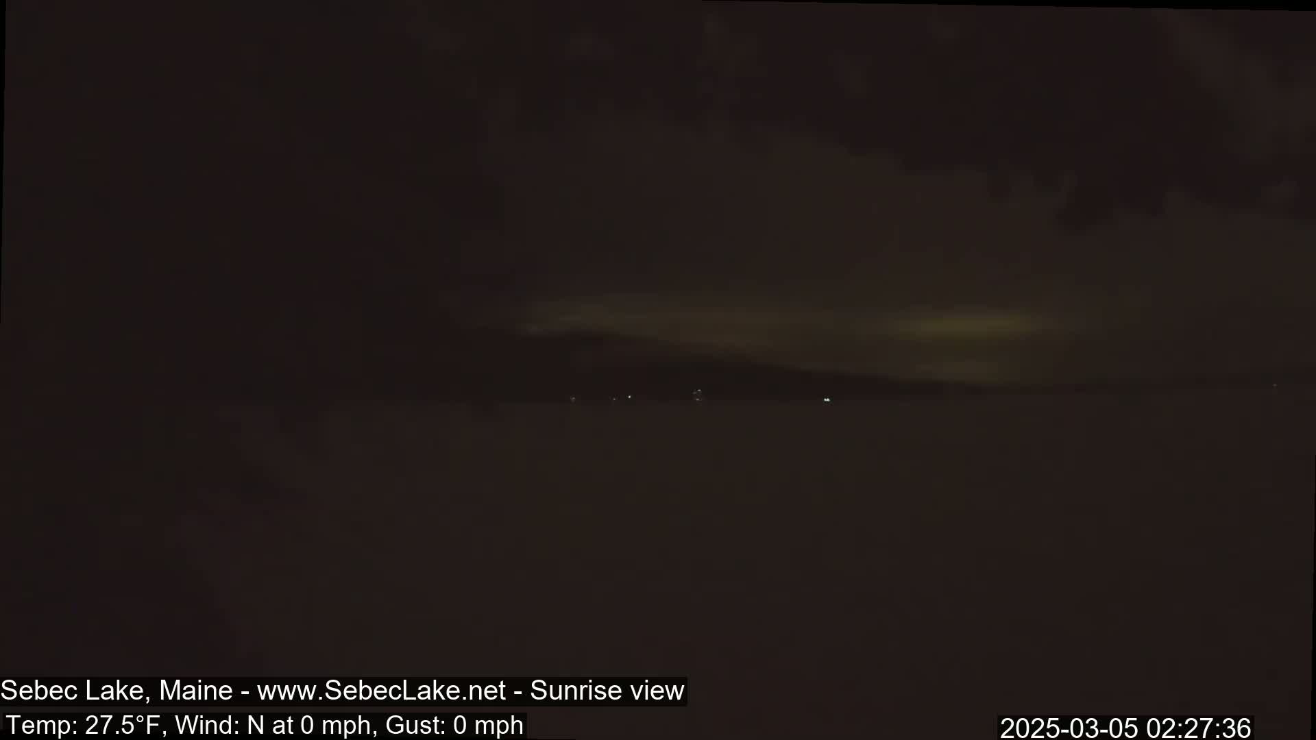 A dark, nighttime view of a calm lake with a faintly illuminated horizon under a mostly cloudy sky.