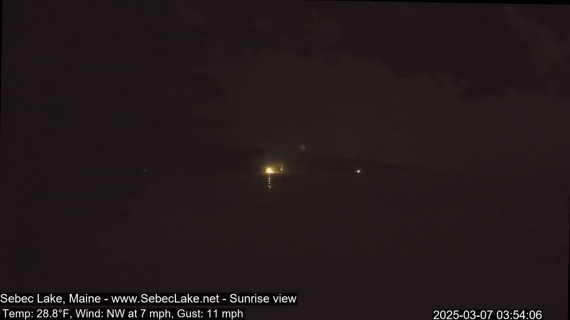 A dark, nighttime scene shows a few faint lights in the distance under a mostly clear sky with a northwest wind of 7 mph and gusts up to 11 mph.