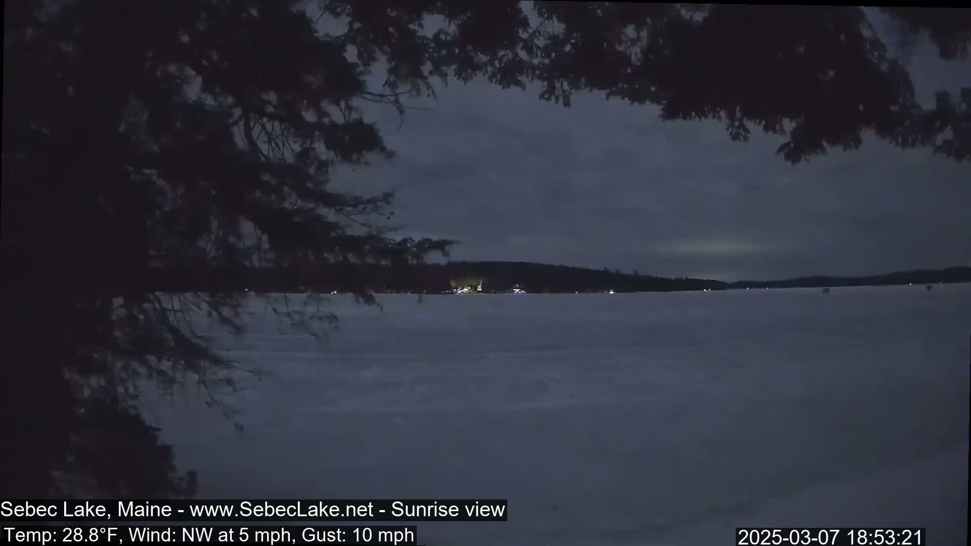 A frozen lake is seen at dusk under a dark, cloudy sky, with tree branches partially obscuring the view.